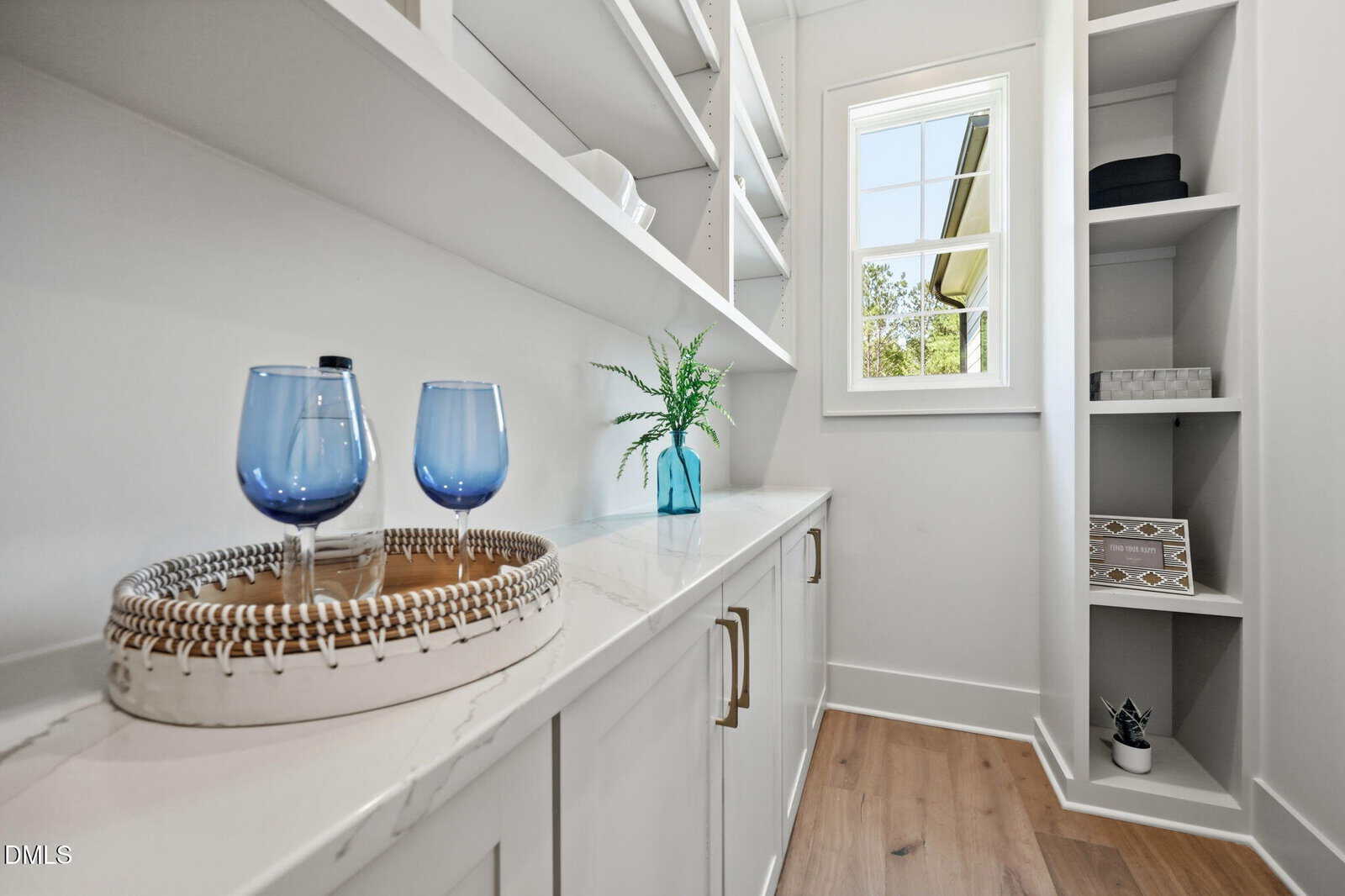 A bright, modern pantry with white cabinets, open shelves, a window, two blue glasses on a tray, a blue vase with greenery, and neatly arranged storage boxes.
