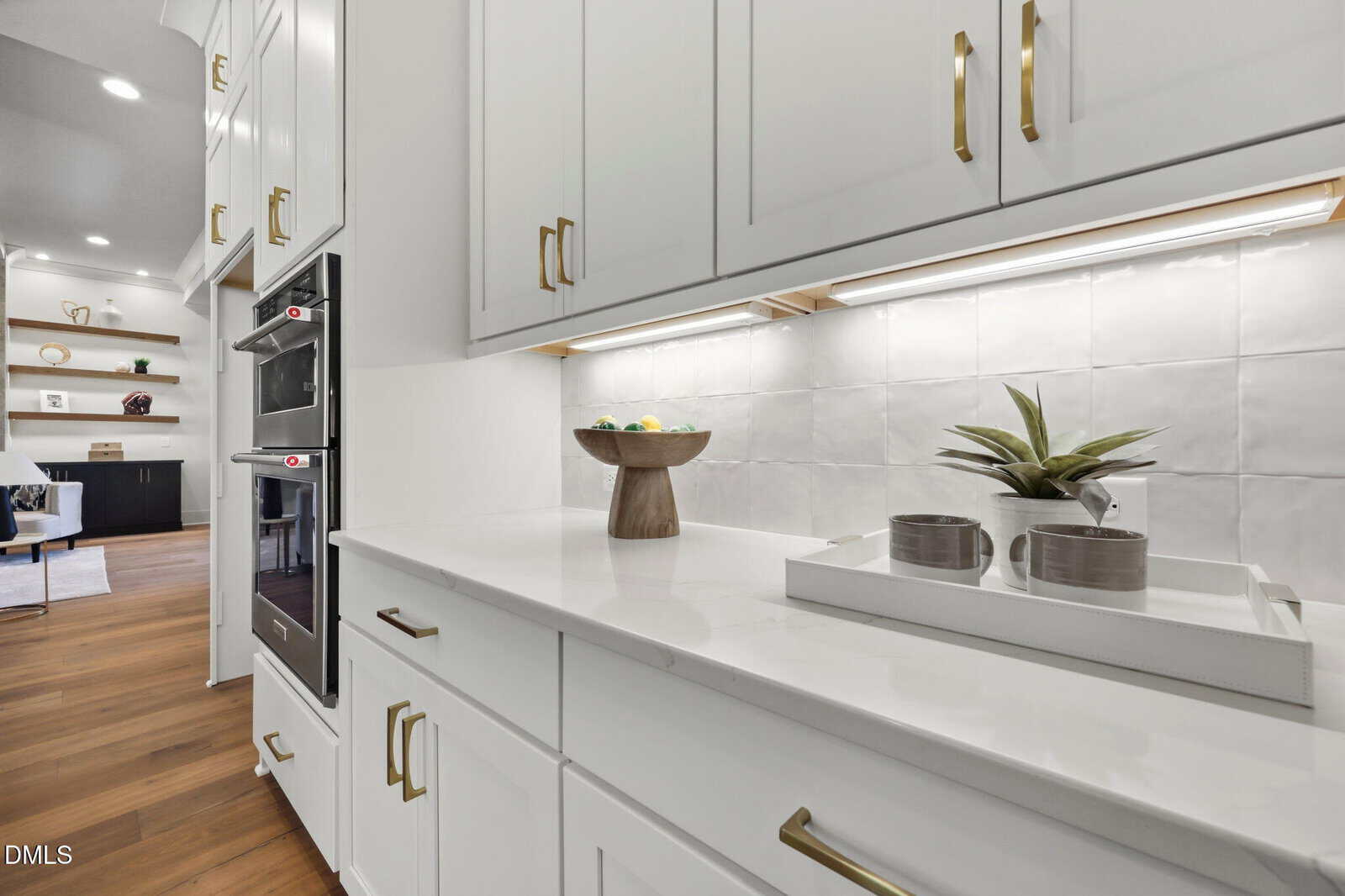 Modern white kitchen with gold hardware, double wall ovens, a countertop holding a wooden fruit bowl, and a white tray with mugs and a small plant. Open shelves with decor are visible in the background.
