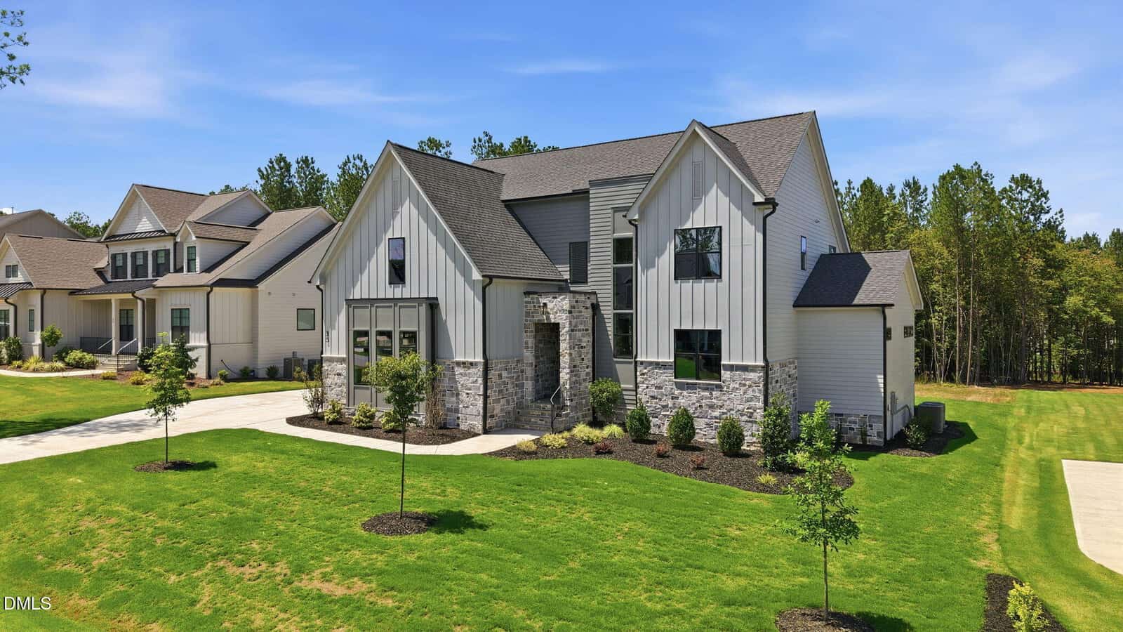 Modern two-story house with white siding, stone accents, and dark trim. Large driveway and manicured lawn with young trees. Neighboring homes and trees in the background under a clear blue sky.