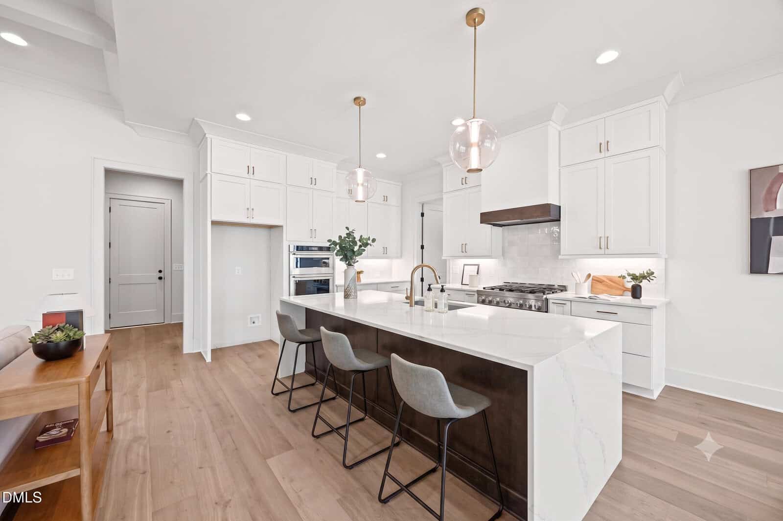 Modern white kitchen with a large marble island, three gray bar stools, pendant lights, built-in ovens, and light wood floors. Minimalist decor with plants and framed art on the side table.