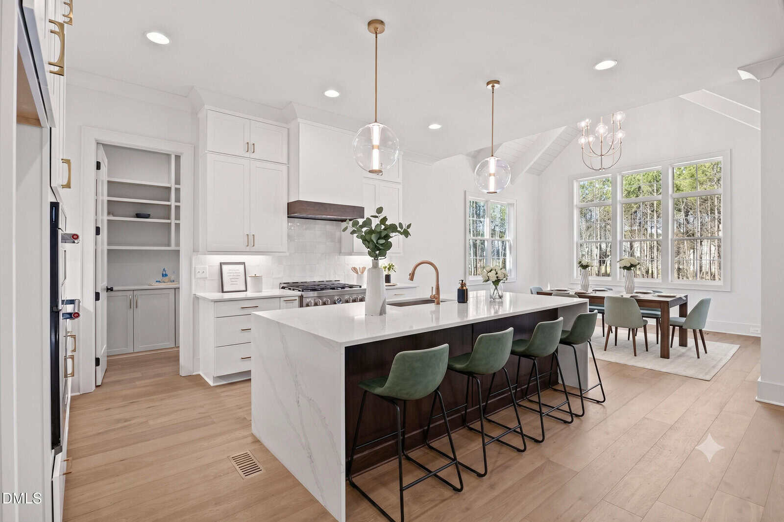 Modern white kitchen with large marble island, green bar stools, gold fixtures, pendant lights, built-in pantry, and adjacent dining area with large windows letting in natural light.