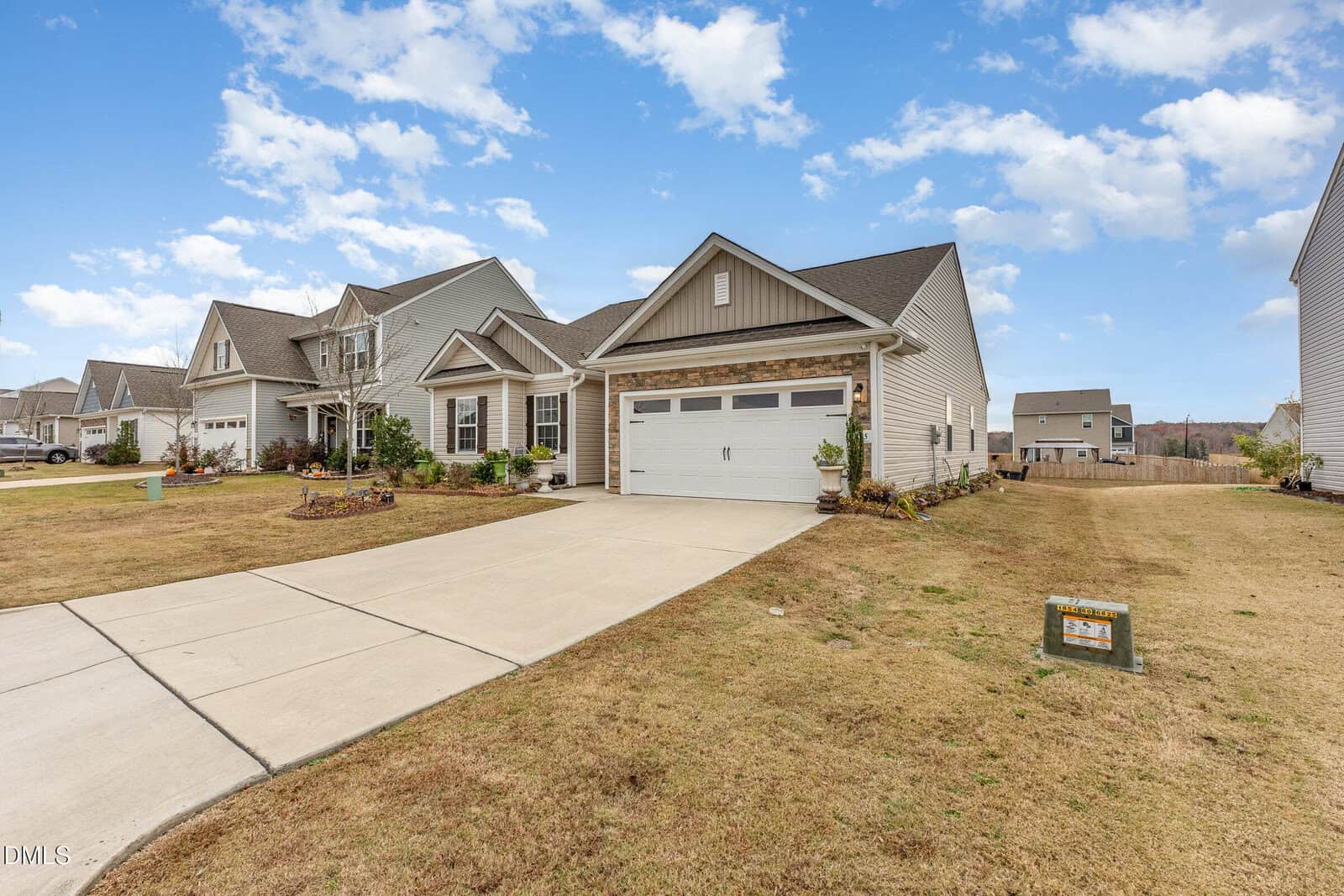Single-story suburban house with a two-car garage, light-colored siding, and stone accents. Concrete driveway and well-kept lawn, with a for sale sign near the street. Other houses and blue sky with clouds in the background.
