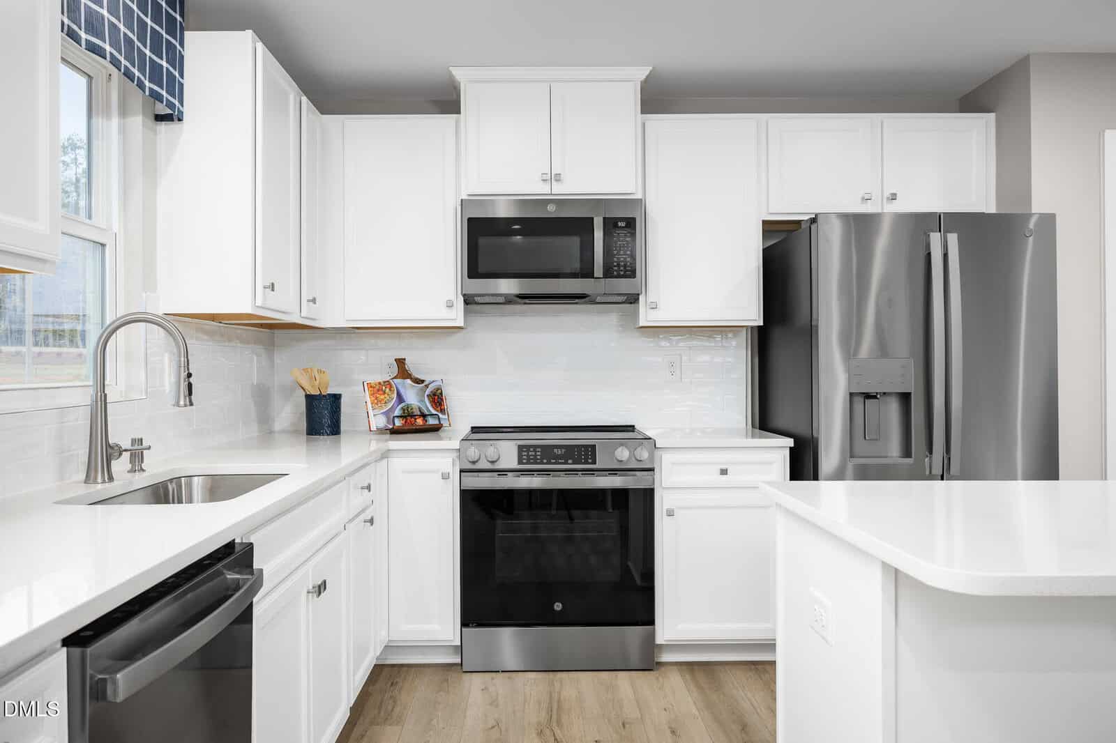 Modern kitchen with white cabinets, stainless steel appliances including a stove, microwave, refrigerator, and dishwasher, white countertops, a window with a blue plaid valance, and wood flooring.