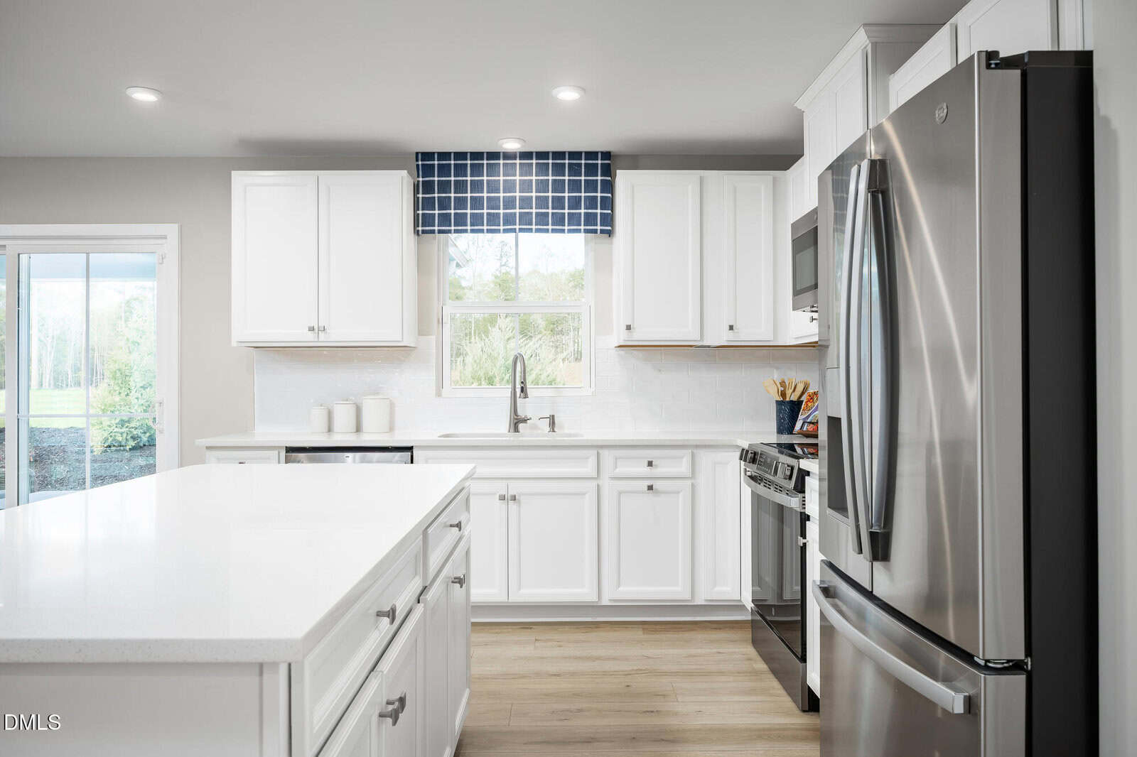 Modern kitchen with white cabinets, stainless steel appliances, a central island, light wood flooring, and a window with a blue checkered valance above the sink. Natural light brightens the space.