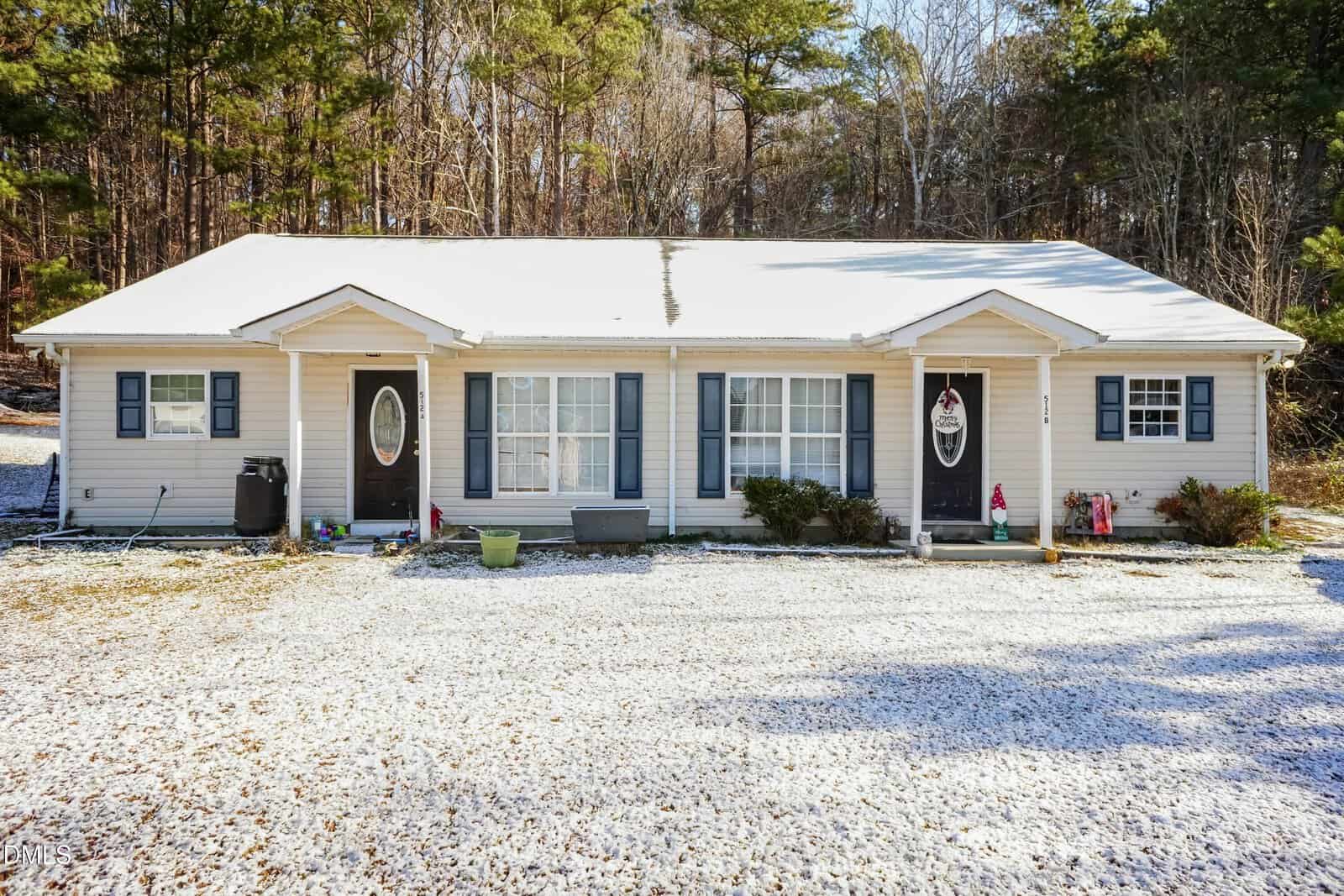 A single-story beige duplex at 512 Cotton Avenue in Creedmoor features two front doors, blue shutters, and a light dusting of snow on the roof and ground. The house is surrounded by trees with minimal landscaping in front.