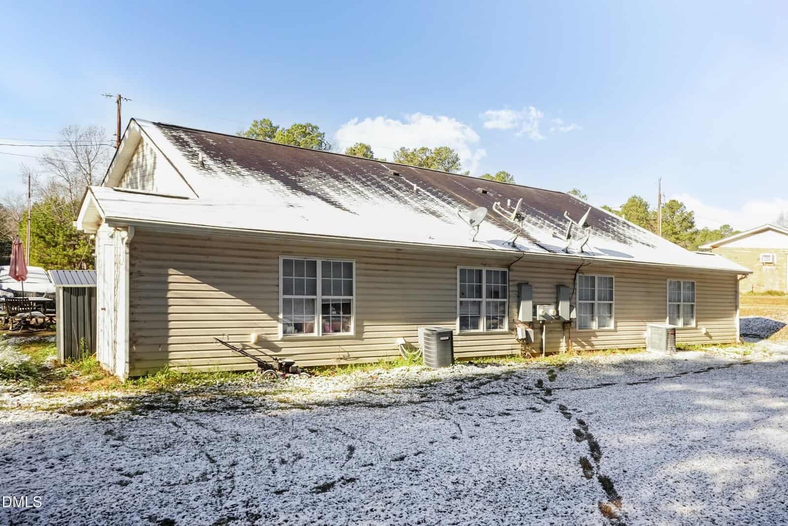 A single-story beige house with a dark roof partially covered in snow. The ground is also dusted with snow, and there are two large windows, air conditioning units, and utility meters along the side. Trees are in the background.