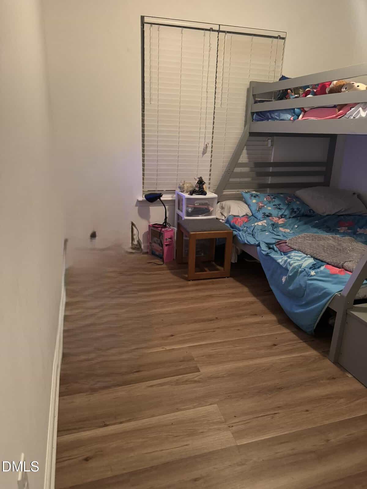A small bedroom with wood flooring, a bunk bed with blue bedding, a window with closed blinds, a small nightstand, and a few toys and storage items near the window. There is a spot on the left wall near the floor.
