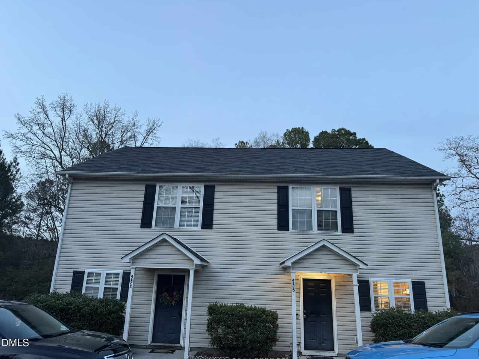 Two-story duplex at 412 W Hillsboro Street in Creedmoor with beige siding, black shutters, and two black front doors. Each unit has a covered entryway and bushes in front. Two cars are partially visible below, with trees in the background.