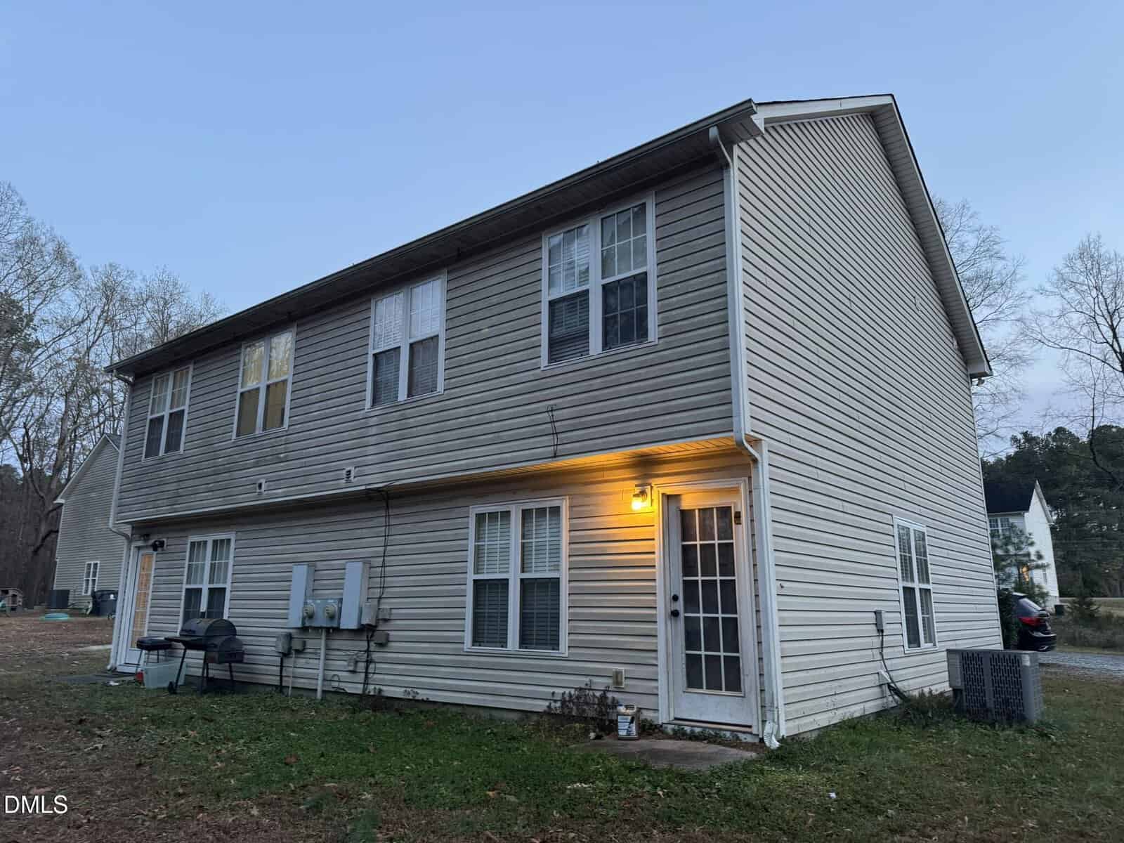 A two-story beige duplex with vinyl siding, multiple windows, a lit porch light by the back door, outdoor utility meters, and a small grill beside the building. The yard is grassy with scattered trees in the background.