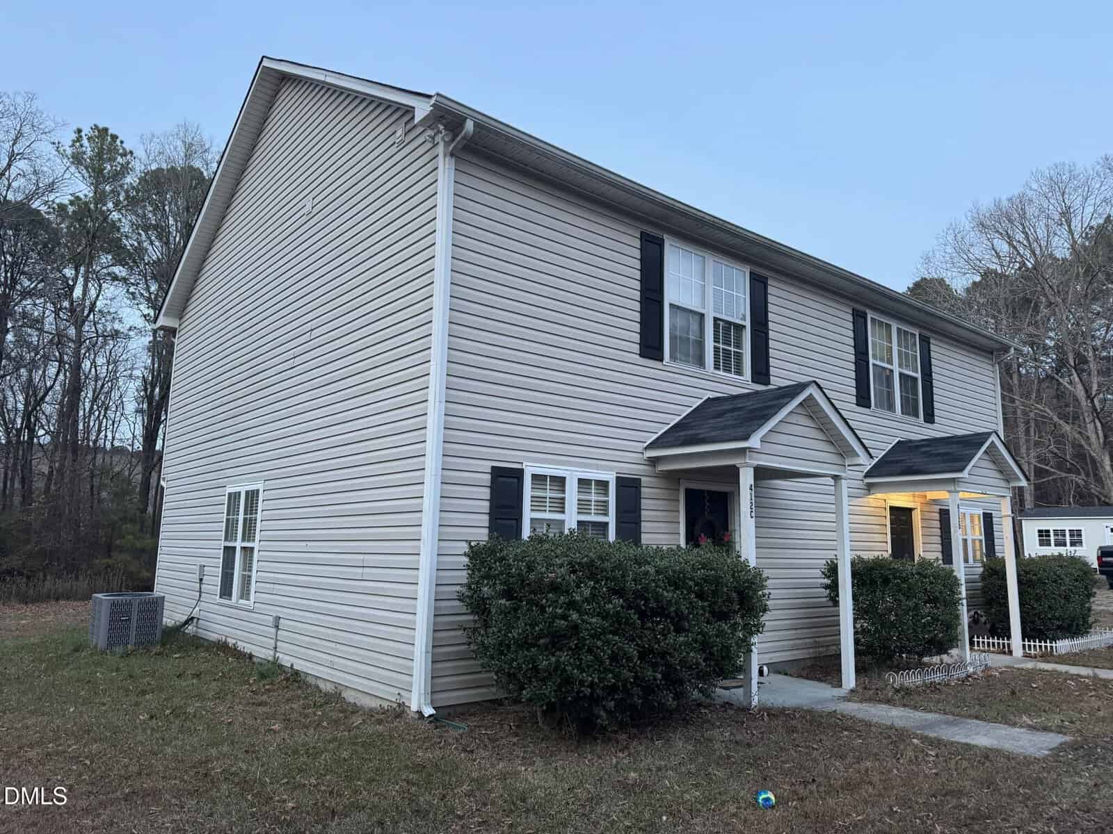 A two-story beige townhouse with black shutters and two separate entrances, each with a small porch and bushes in front. The yard is grassy with trees and an air conditioning unit on the side.