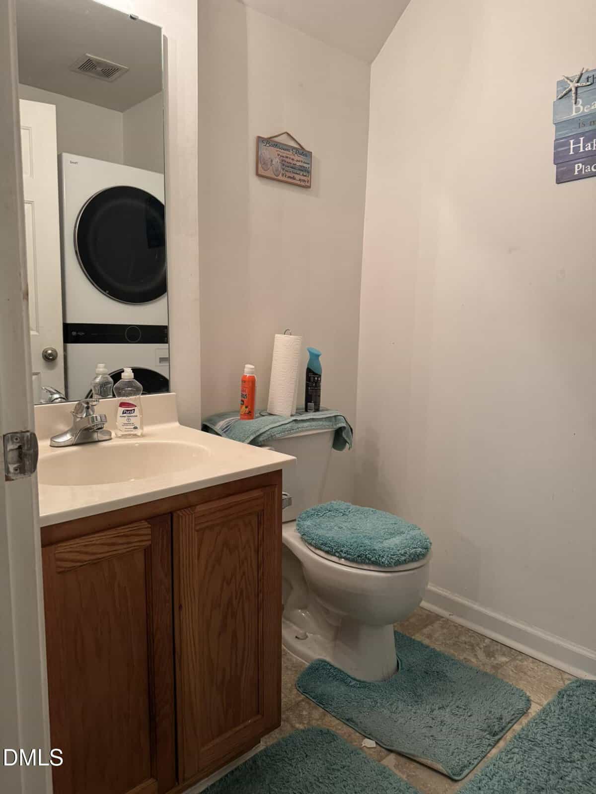 A small bathroom with a wooden cabinet sink, a toilet with teal rugs, and a teal bathmat. Cleaning supplies and paper towels are on the toilet tank. A stacked washer and dryer are visible in the mirror.