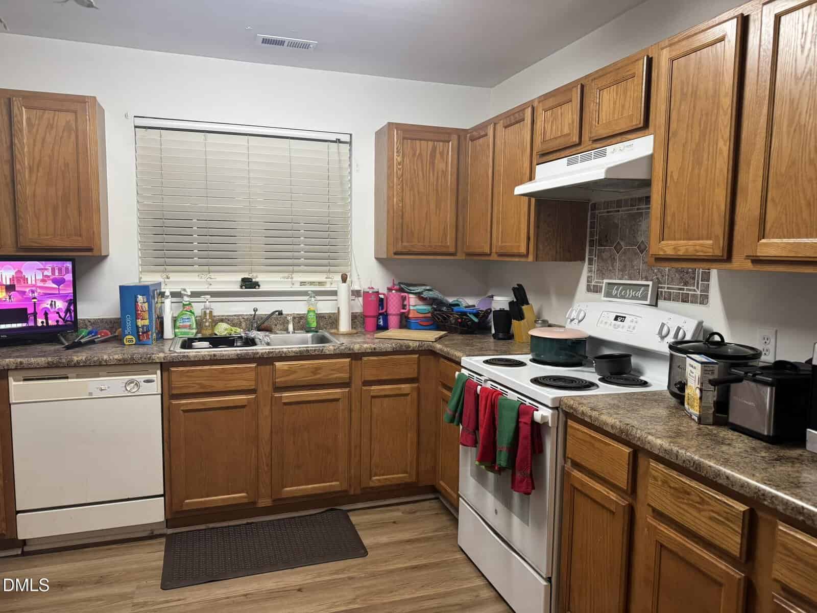 A kitchen with brown wooden cabinets, white appliances including a stove and dishwasher, a countertop with various items, a double sink under a window with closed blinds, and a television on the left side.