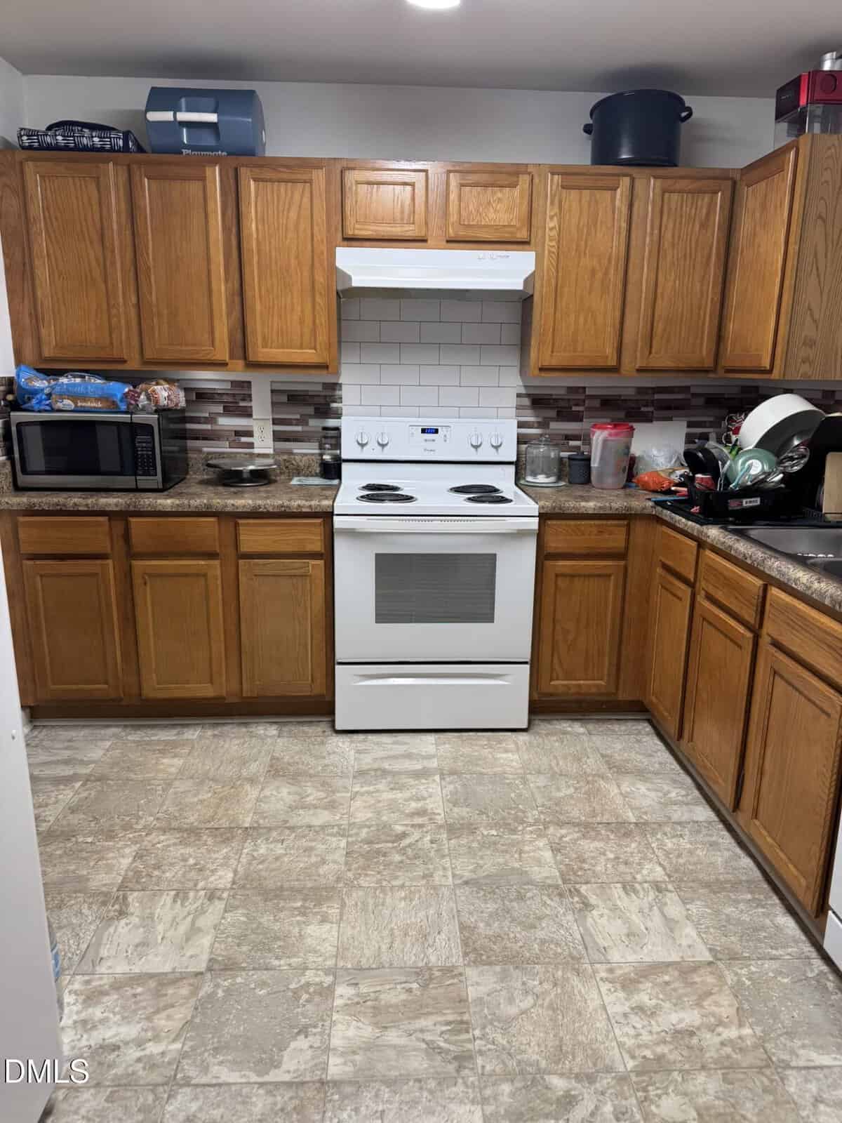 A kitchen with brown wooden cabinets, a white electric stove and oven, a microwave, dishes on a drying rack, and various kitchen items on the countertops. The backsplash is tiled, and the floor has a beige pattern.