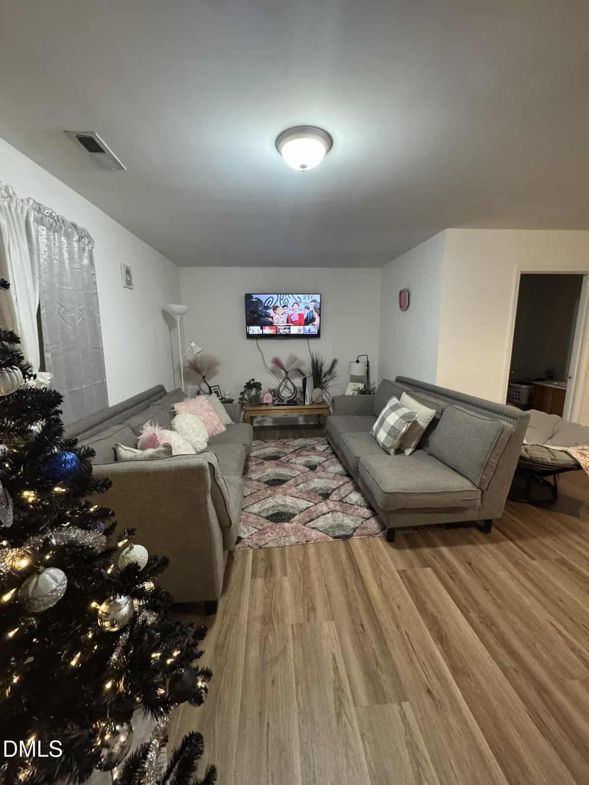 A living room with two gray sofas, a TV mounted on the wall, a coffee table, a decorated Christmas tree, wood-look flooring, and a rug with a geometric pattern. The TV displays a news program.