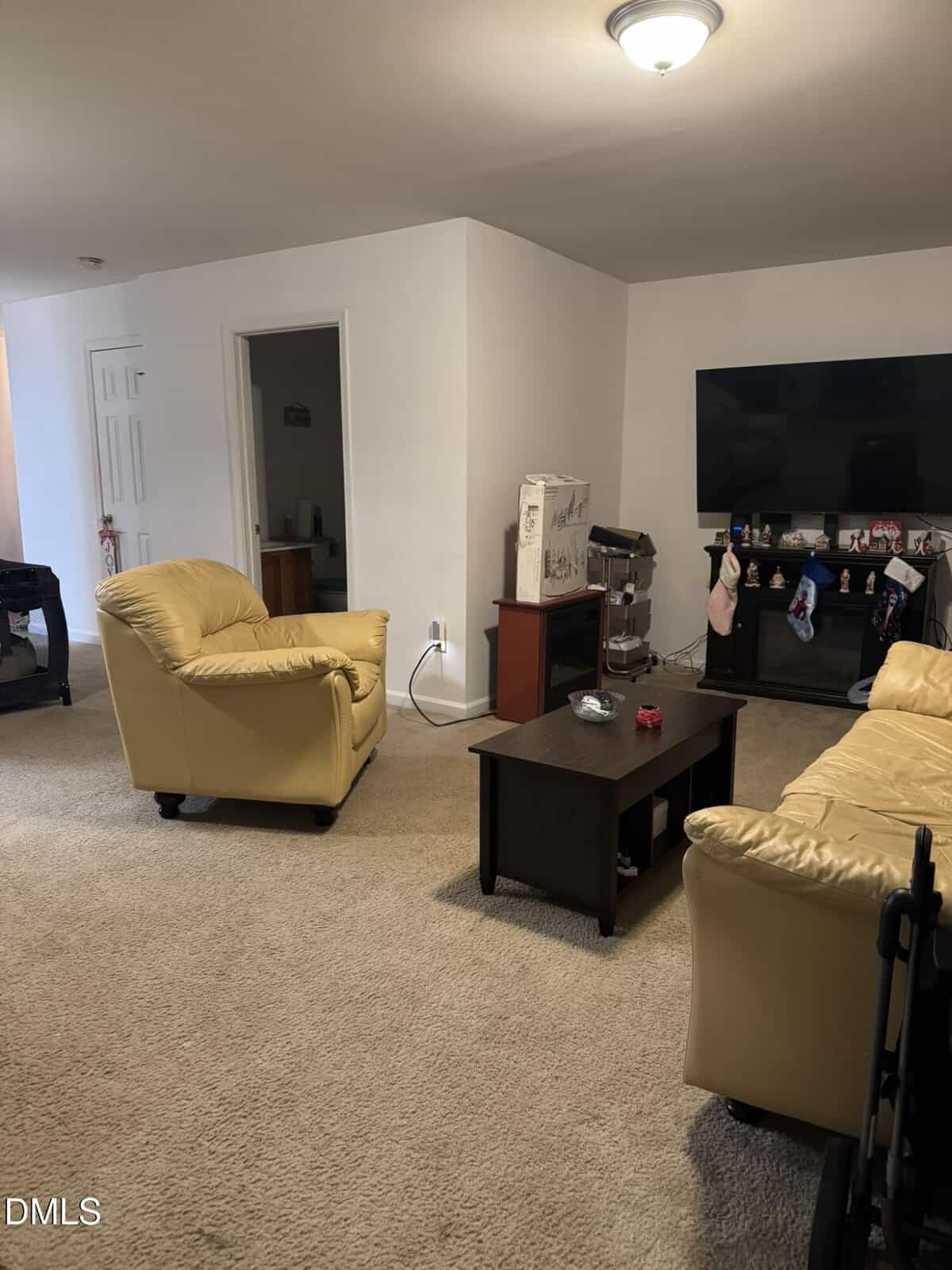 A living room with tan carpet, yellow armchairs, a matching sofa, a dark coffee table, a TV on a stand, and assorted items on the TV stand and coffee table. A doorway to another room is visible in the background.