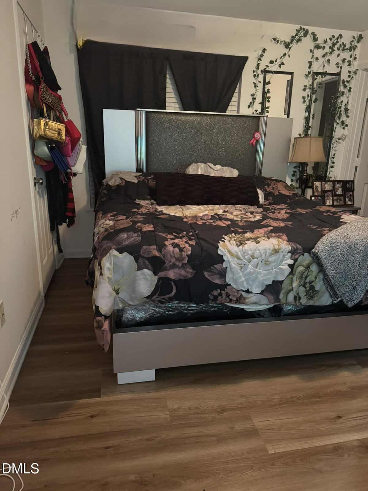 A bedroom with a large bed covered in a floral-patterned comforter, wood flooring, a nightstand with a lamp, hanging ivy decor, purses and hats on the wall, and dark curtains over the window.