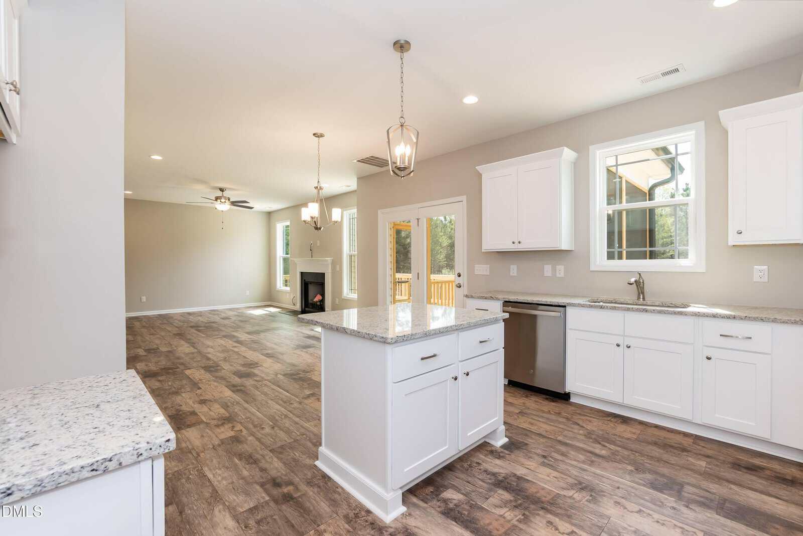 A modern kitchen with white cabinets and granite countertops, open to a spacious living area with wood floors, large windows, and a door leading to a deck outside. Pendant lights hang above the kitchen island.
