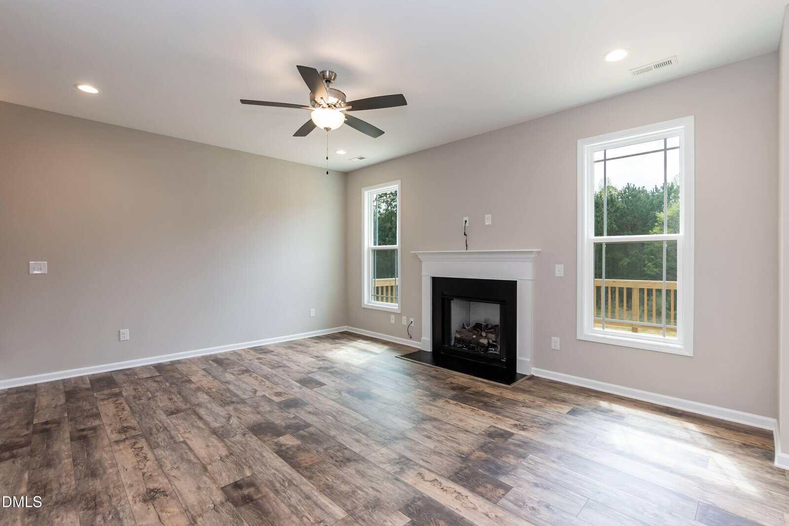 A spacious, empty living room with wood flooring, light gray walls, a ceiling fan with lights, a white fireplace, and two large windows letting in natural light.