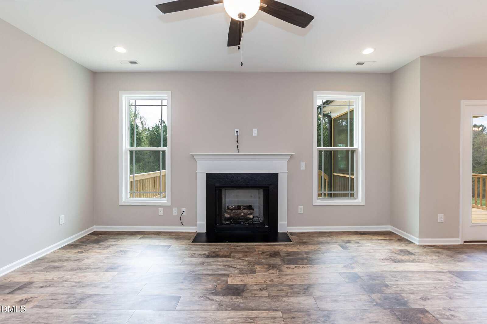 Bright living room with wood-look flooring, a central black fireplace, light gray walls, and two tall windows on either side letting in natural light. Ceiling fan with a light fixture is above.