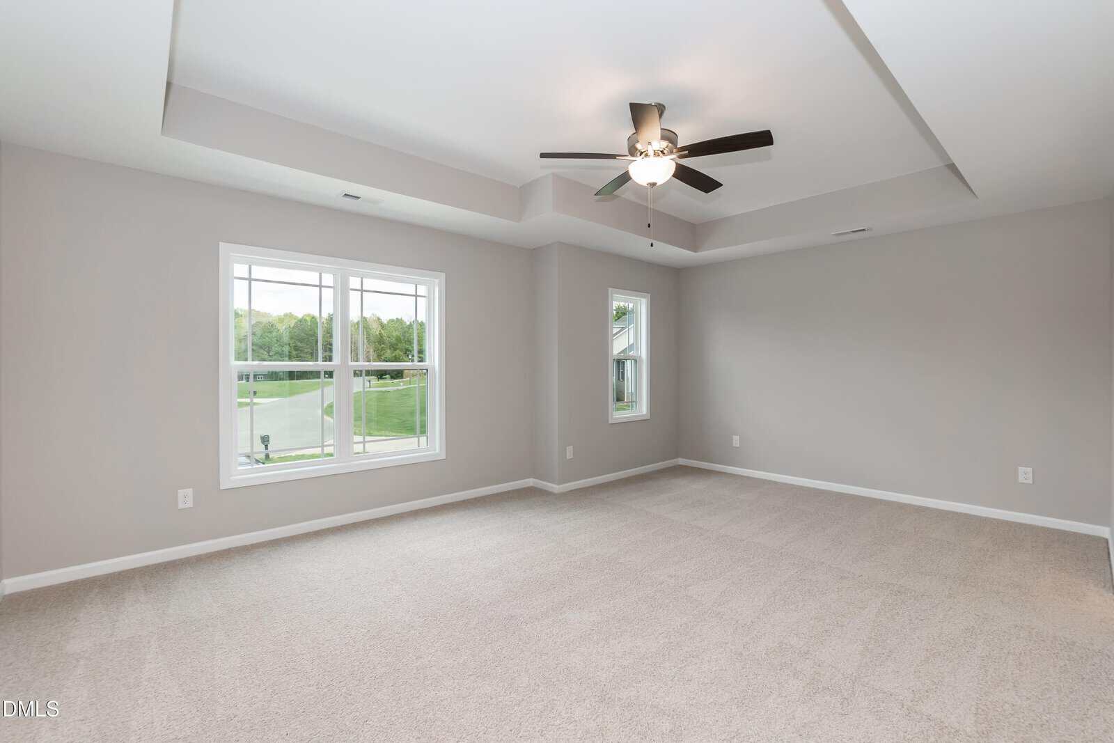 A spacious, empty room with light beige carpet, gray walls, two large windows, a tray ceiling, and a ceiling fan with lights. Natural light streams in through the windows, revealing a green view outside.