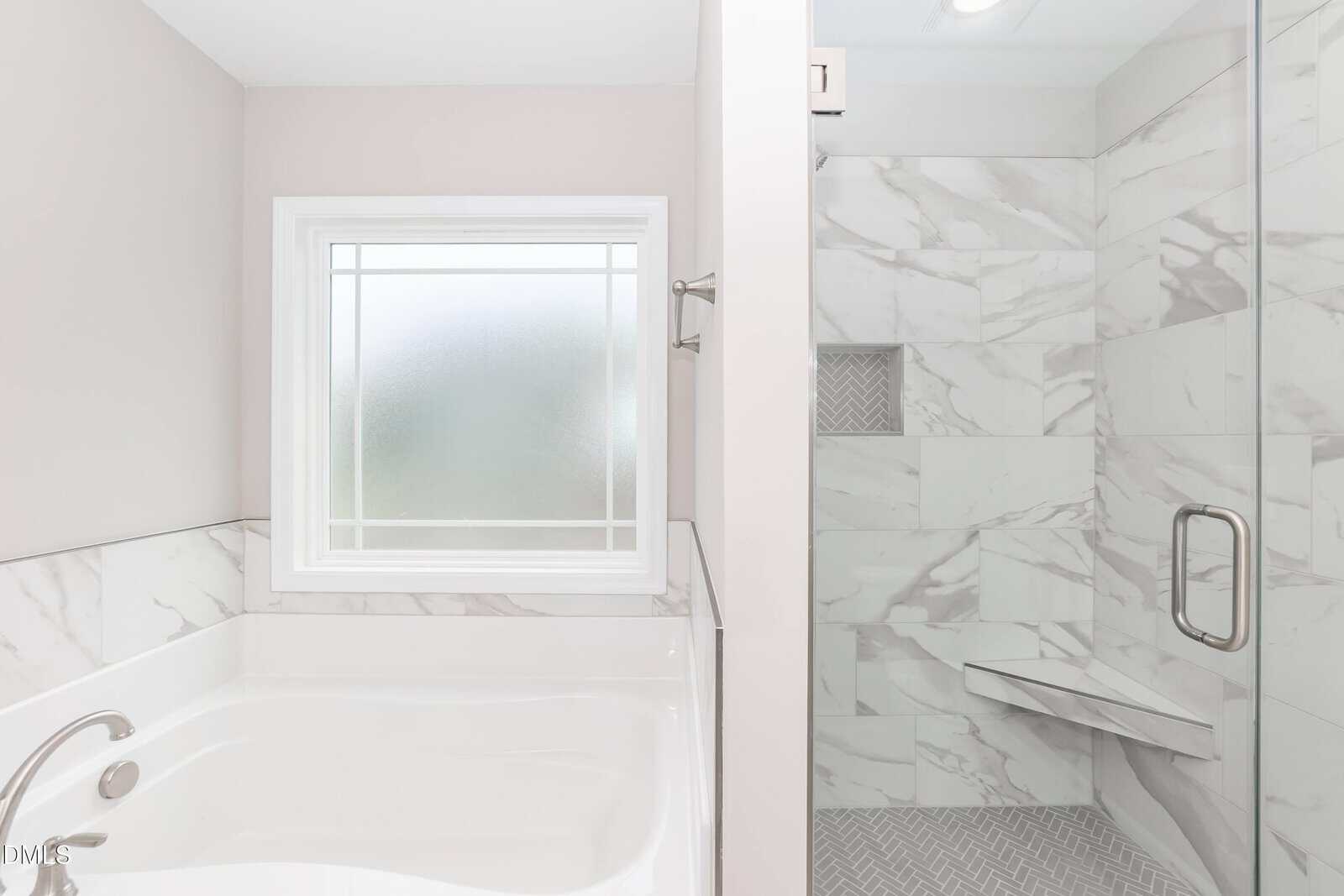 Modern bathroom with a white bathtub below a frosted window, next to a glass-enclosed shower featuring a built-in bench and marble-patterned tiles on the walls and floor.