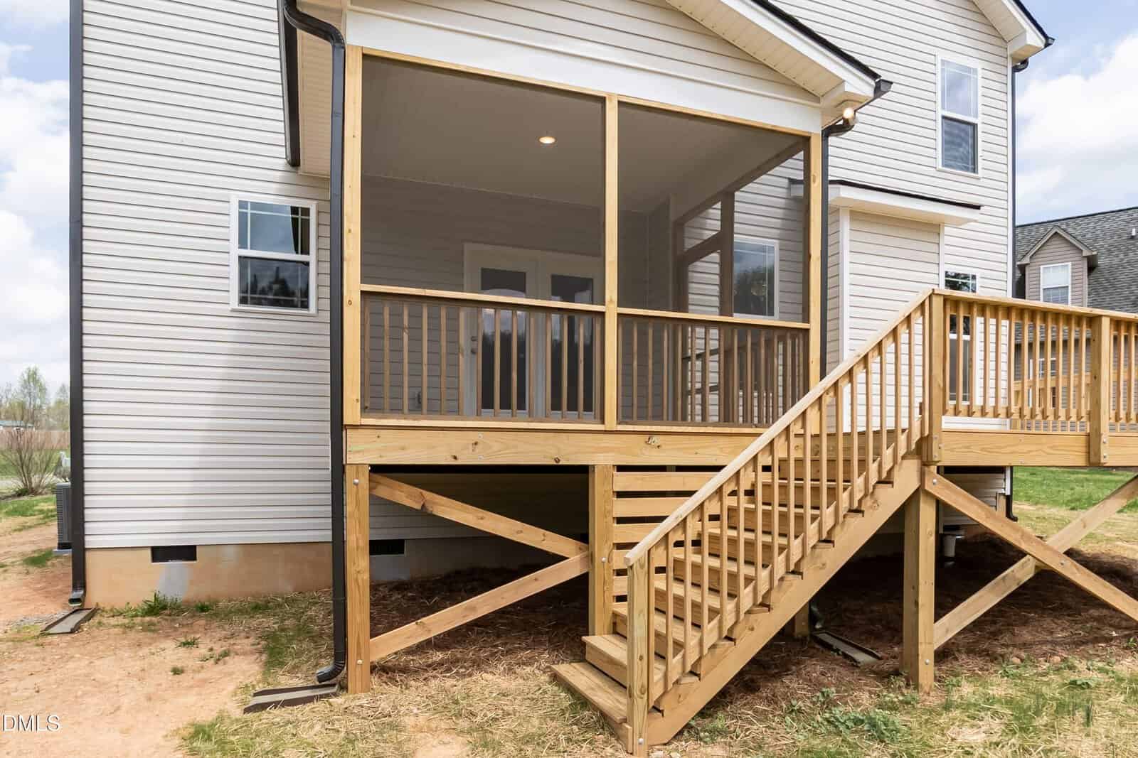 A wooden staircase leads up to a screened-in porch at the back of a beige house with white trim. The porch has wooden railings and sits above a patchy grass and dirt yard.