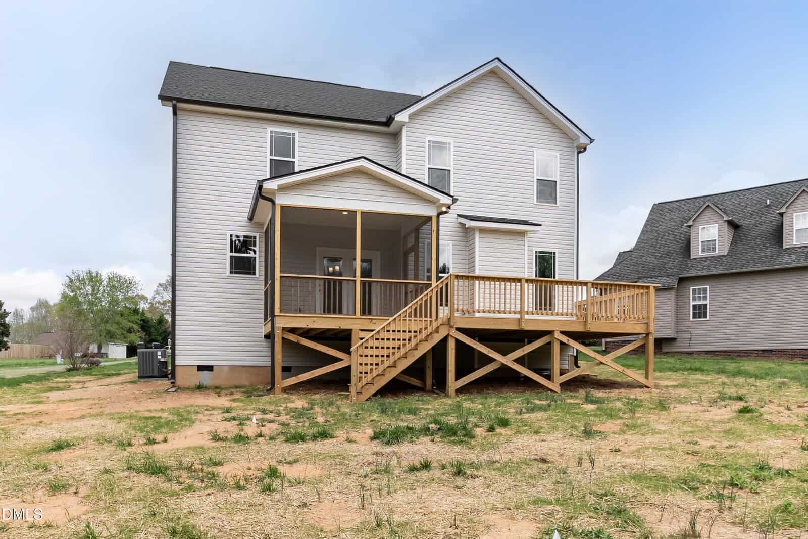 Two-story house with light-colored siding and a dark roof, featuring a large wooden deck and stairs leading to a spacious, partially grassy backyard. Another house is visible to the right.