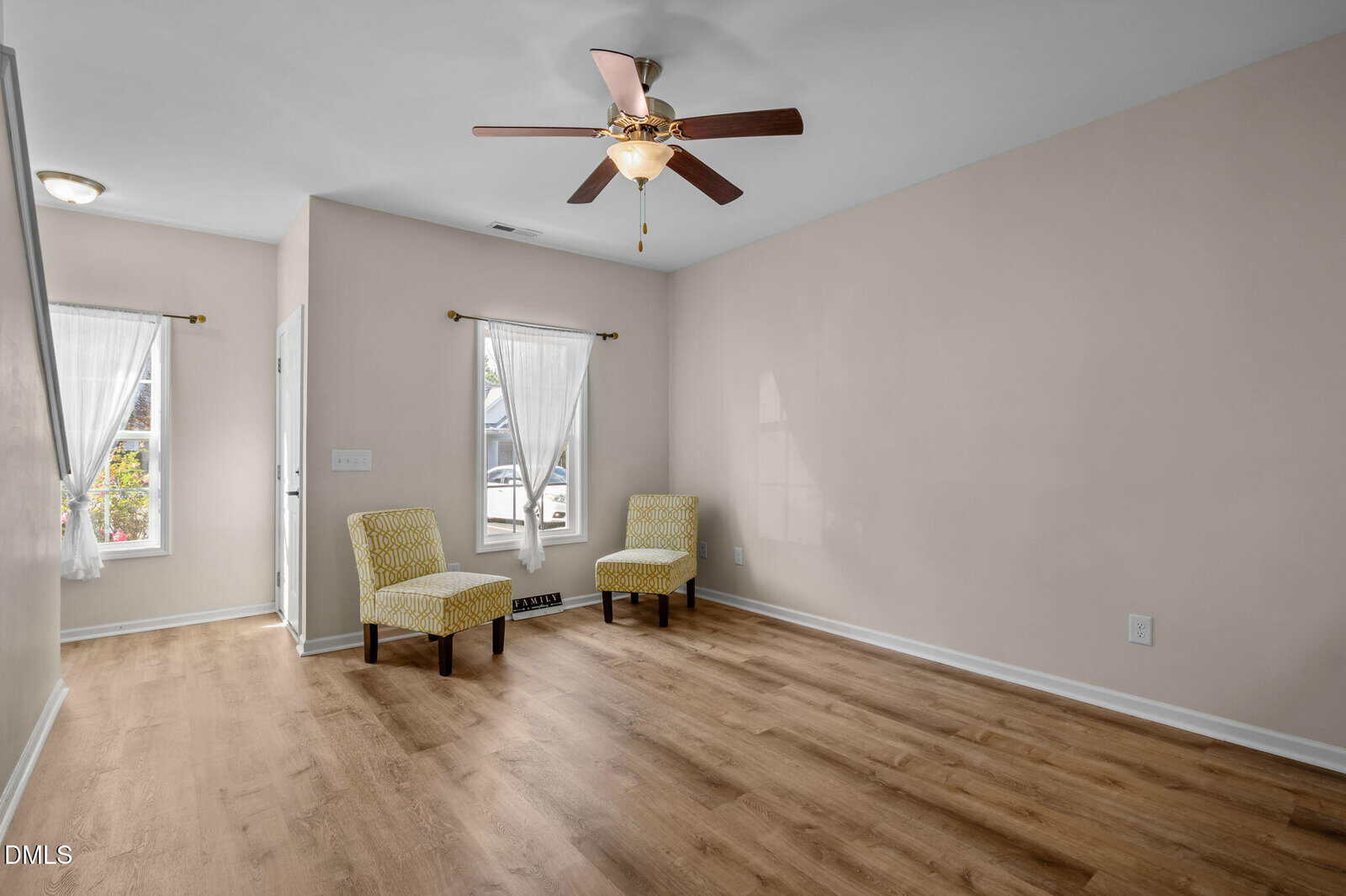 A mostly empty living room with light wood flooring, beige walls, two windows with sheer white curtains, two patterned yellow chairs, and a ceiling fan with a light.