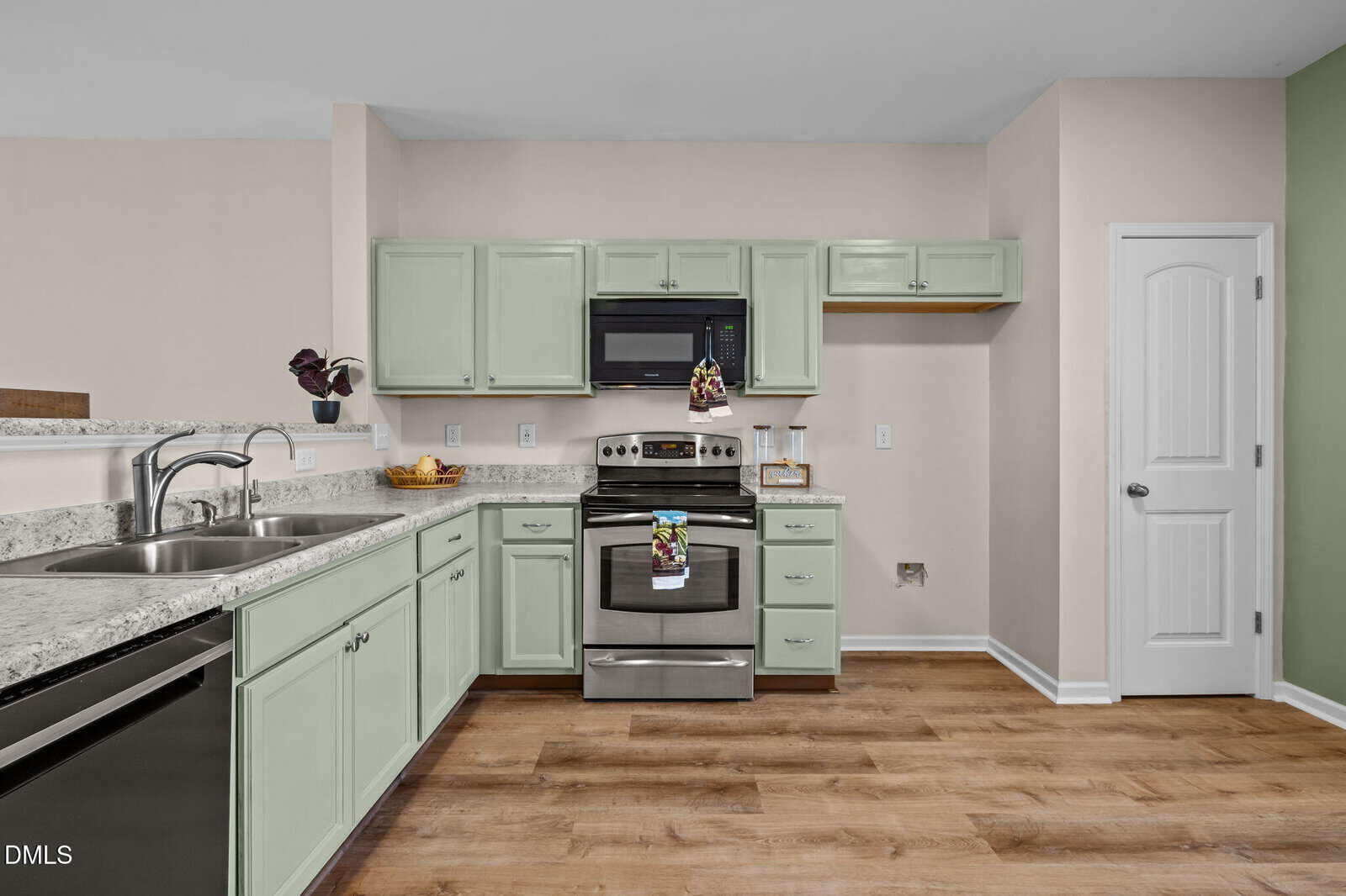 A kitchen with light green cabinets, stainless steel appliances, marble countertops, a double sink, light wood flooring, and light pink and green walls. A white door is on the right.