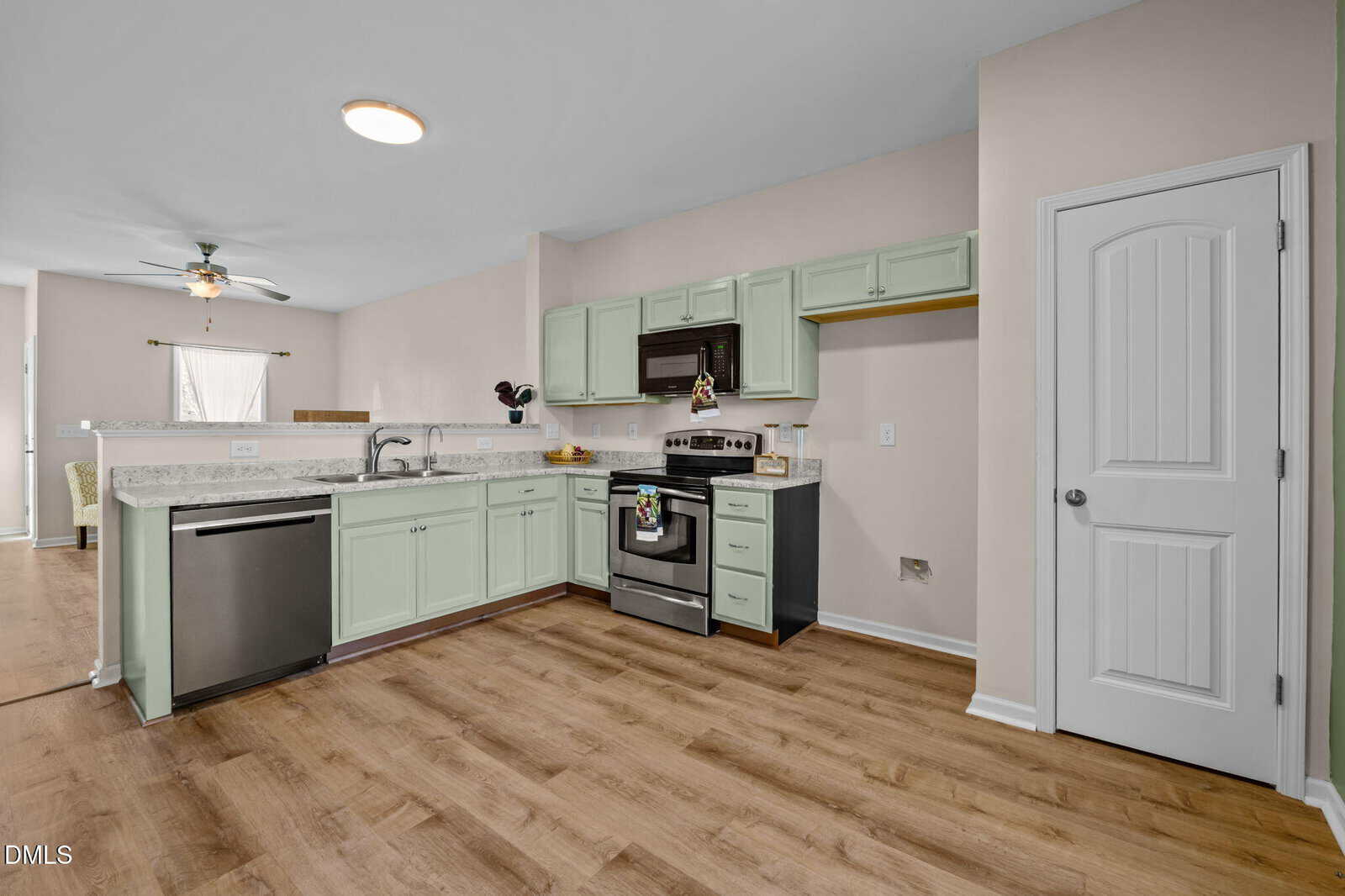 A kitchen with light green cabinets, stainless steel appliances, a double sink, and a marble countertop, featuring wood flooring and a partially open layout to a dining area.