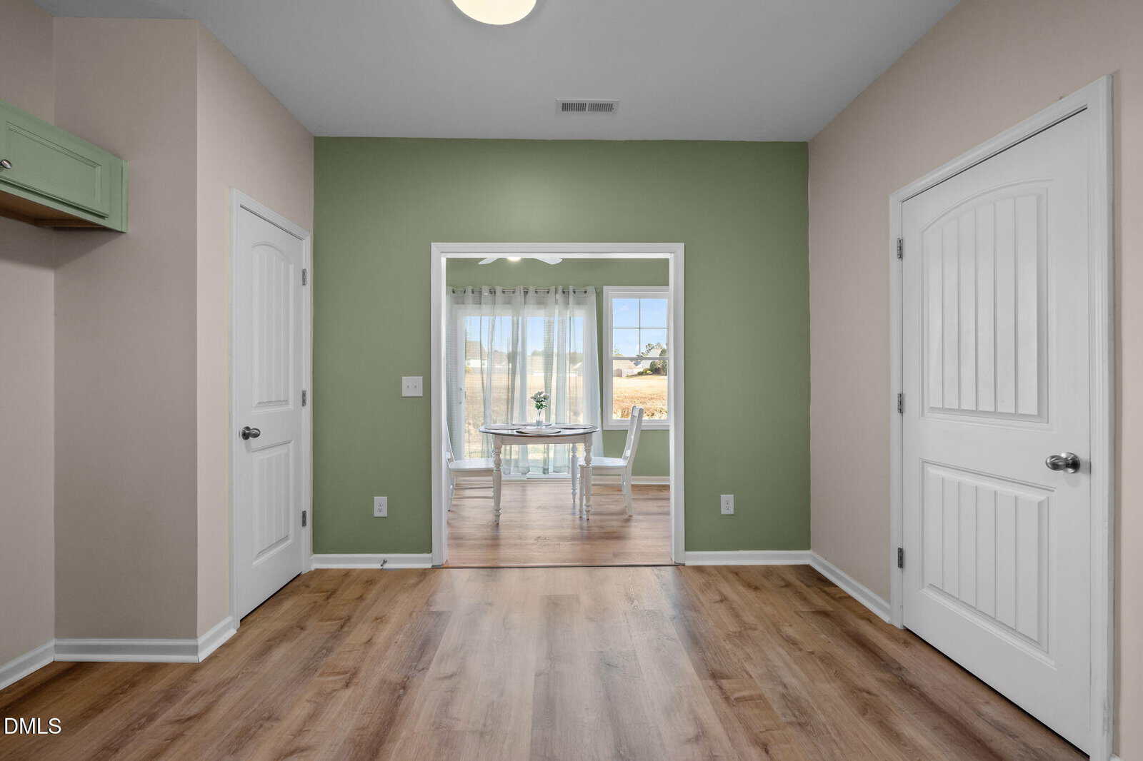 A room with light wood floors, beige and green walls, two white doors, and an open doorway leading to a bright dining area with a white table, chairs, and large windows with sheer curtains.