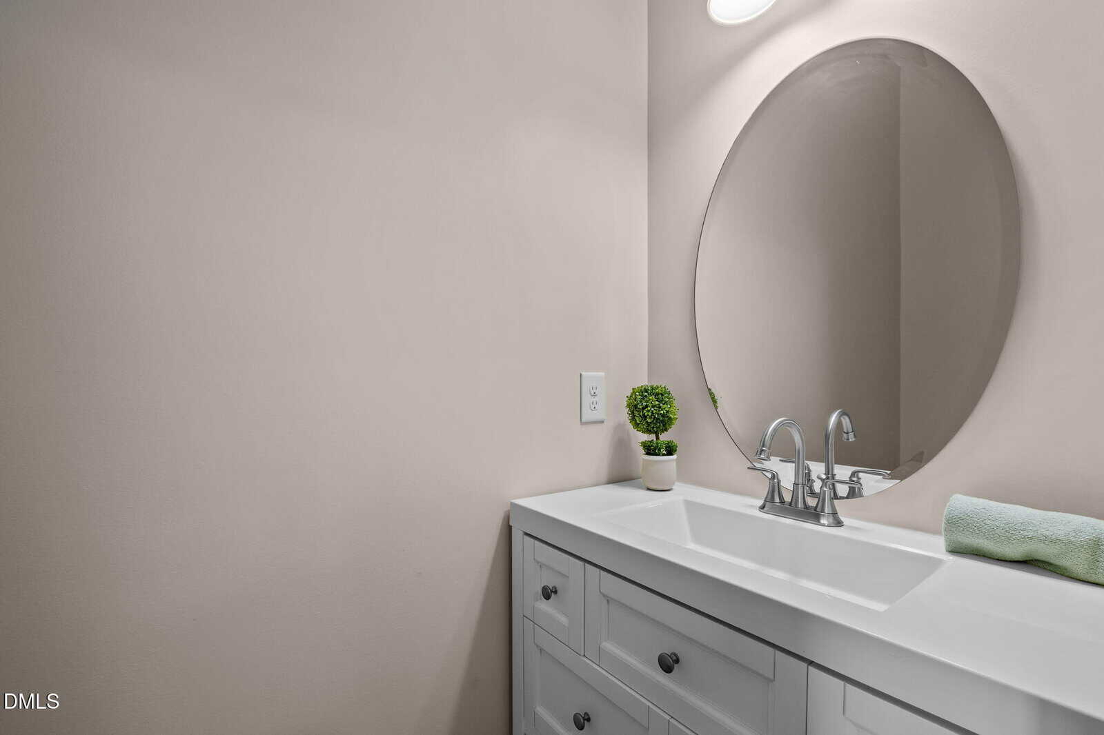 A modern bathroom vanity with a white countertop, oval mirror, chrome faucet, small potted plant, and a rolled green towel against a beige wall.