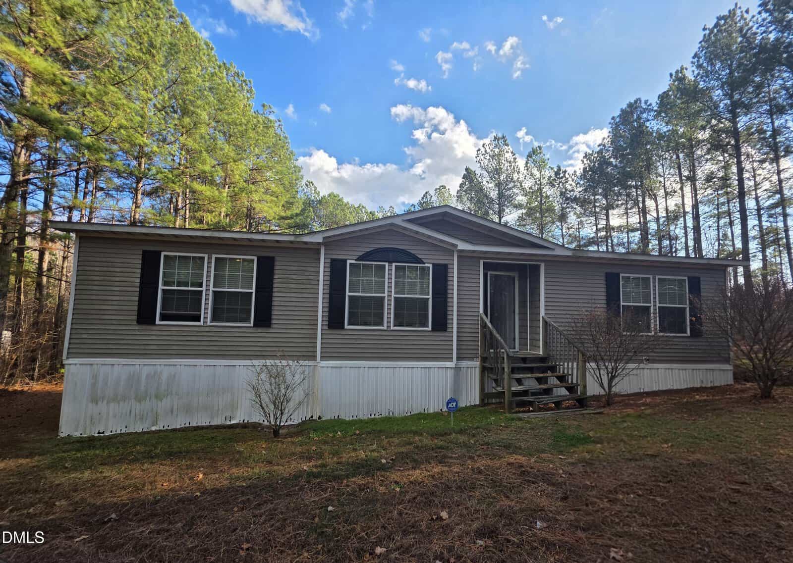 A single-story manufactured home with beige siding, black shutters, and a covered front porch with steps sits at 664 Glebe Road in Henderson, nestled among tall pine trees under a partly cloudy sky.