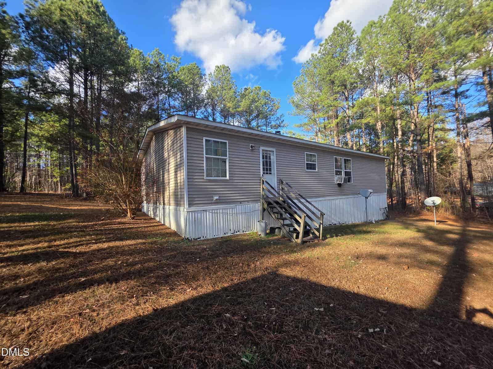 A single-story manufactured home with beige siding and white trim, surrounded by pine trees. Wooden steps lead to the front door. The yard is covered with brown pine needles and two satellite dishes are visible.