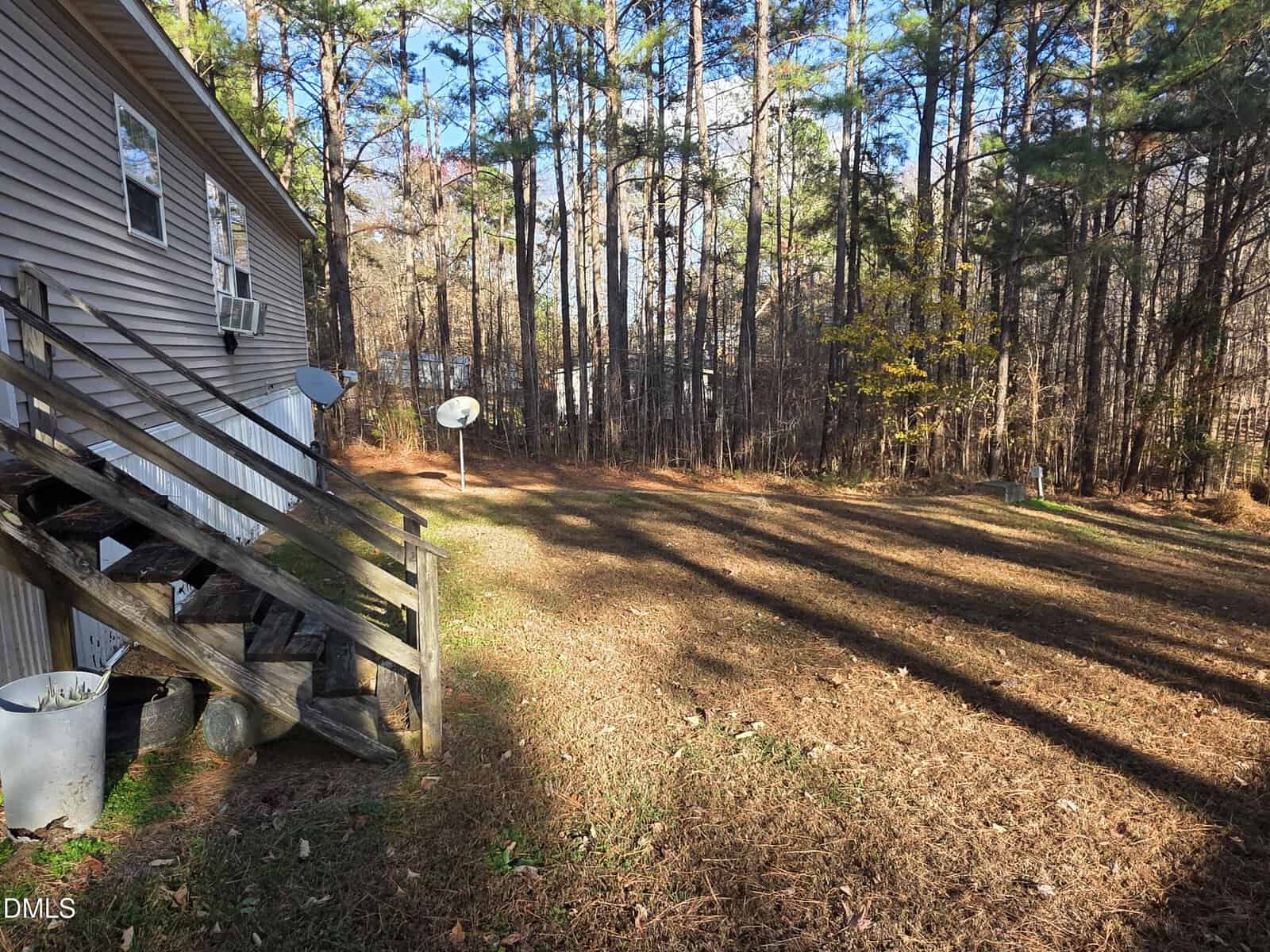 A wooden staircase leads up to the side door of a beige house on the left. The yard is covered in pine needles, bordered by a forest of tall trees. Two satellite dishes are visible near the edge of the woods.