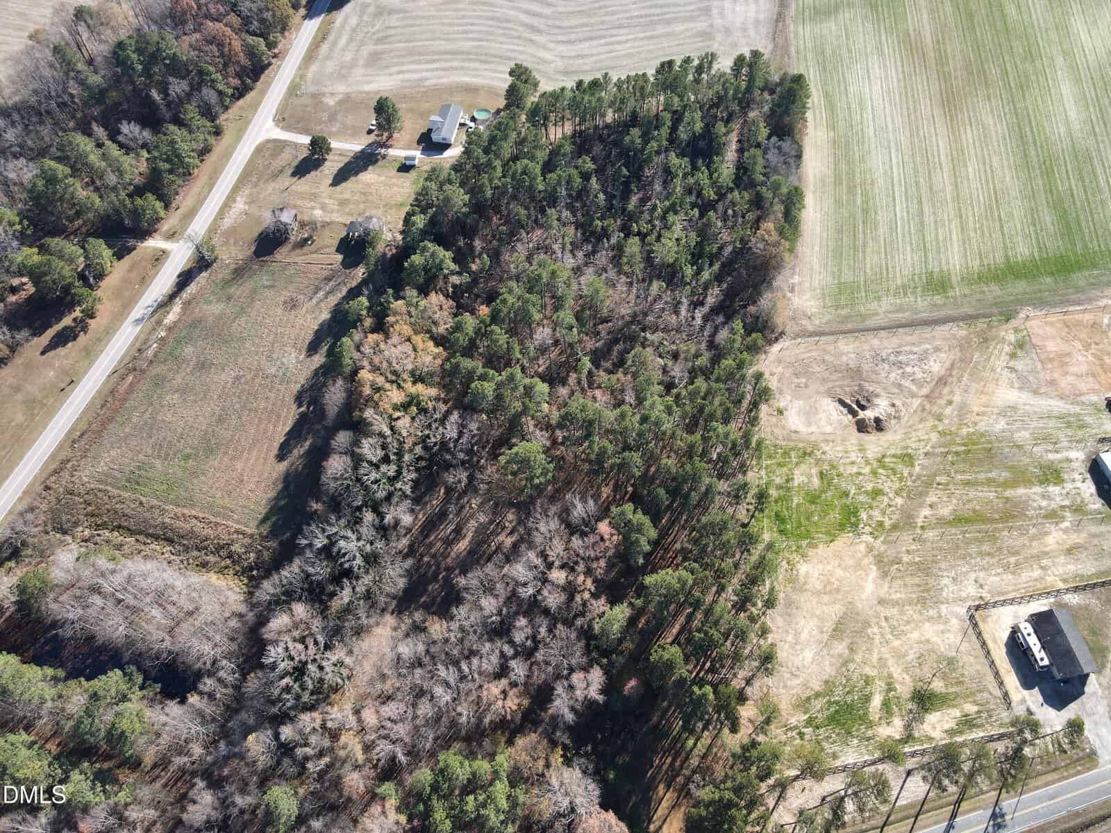 Aerial view of a rural landscape featuring a wooded area, surrounding fields, a small house near a road, and patches of cleared land with visible tire tracks.
