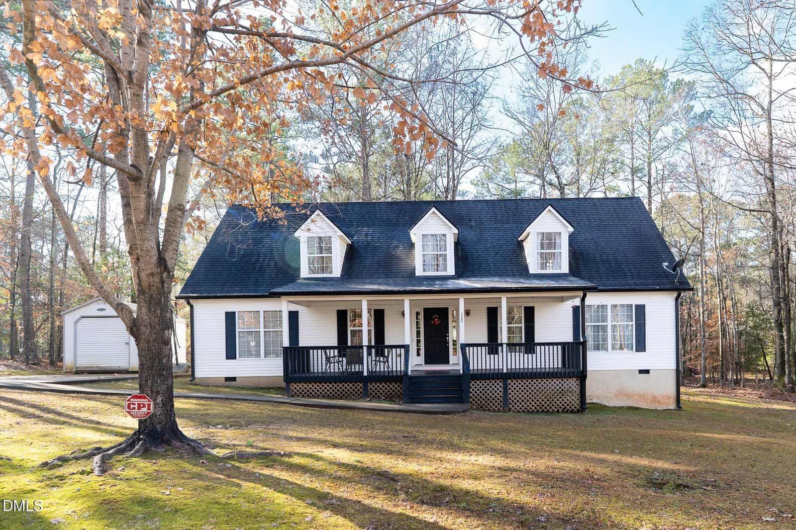 A charming white house with black trim and a covered front porch sits on a wooded yard at 132 Osage Drive in Louisburg, surrounded by autumn leaves. Three dormer windows are on the roof, and a detached garage is visible to the left behind the house.
