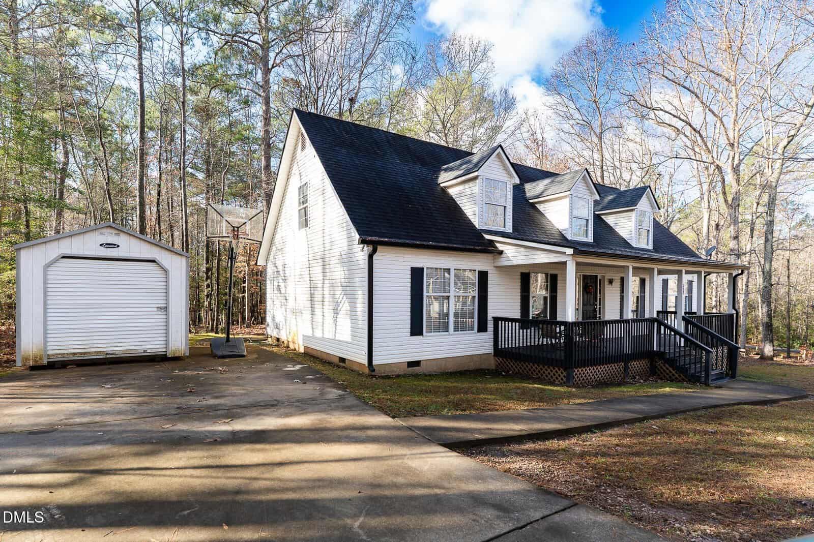 A white, two-story house with black shutters and a covered front porch sits beside a detached single-car garage on a concrete driveway, surrounded by tall trees and a spacious yard.