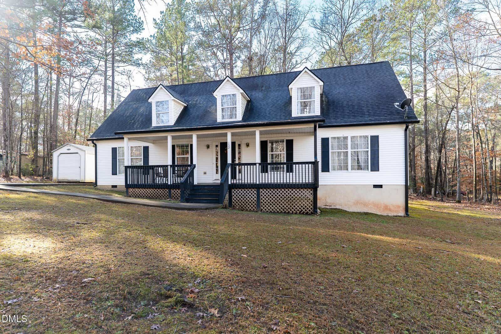 A white house with black trim and a front porch, featuring three dormer windows on the roof. The house sits on a grassy yard surrounded by trees and has a small detached shed in the background.