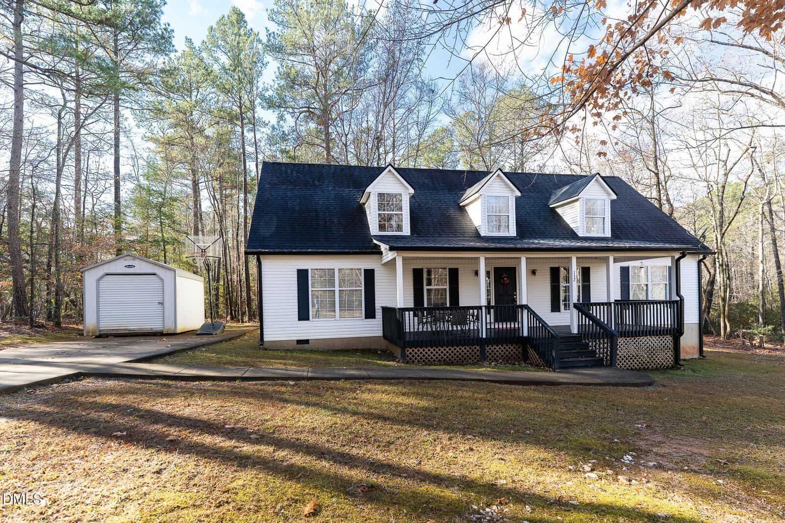 A white two-story house with black shutters and a large front porch, set in a wooded area with a detached garage, surrounded by trees and fallen leaves, under a partly cloudy sky.