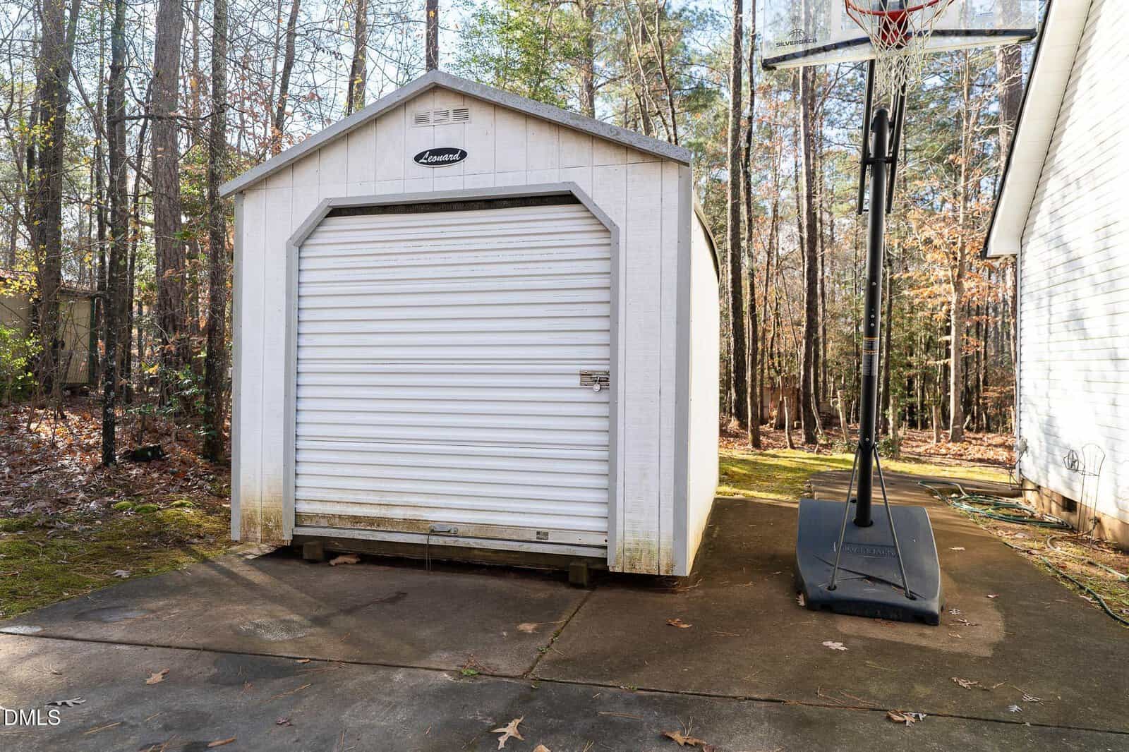 A white metal storage shed with a roll-up door stands on a concrete pad next to a portable basketball hoop, with trees and a house visible in the background.