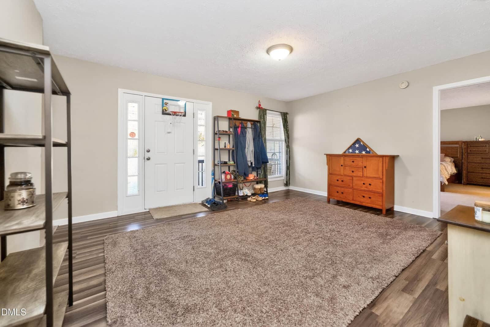 Spacious living room with a brown rug, wooden shelving, a wooden dresser with a folded flag, a coat rack, and a white front door with windows. Light walls and wood flooring, with a bedroom visible through an open doorway.