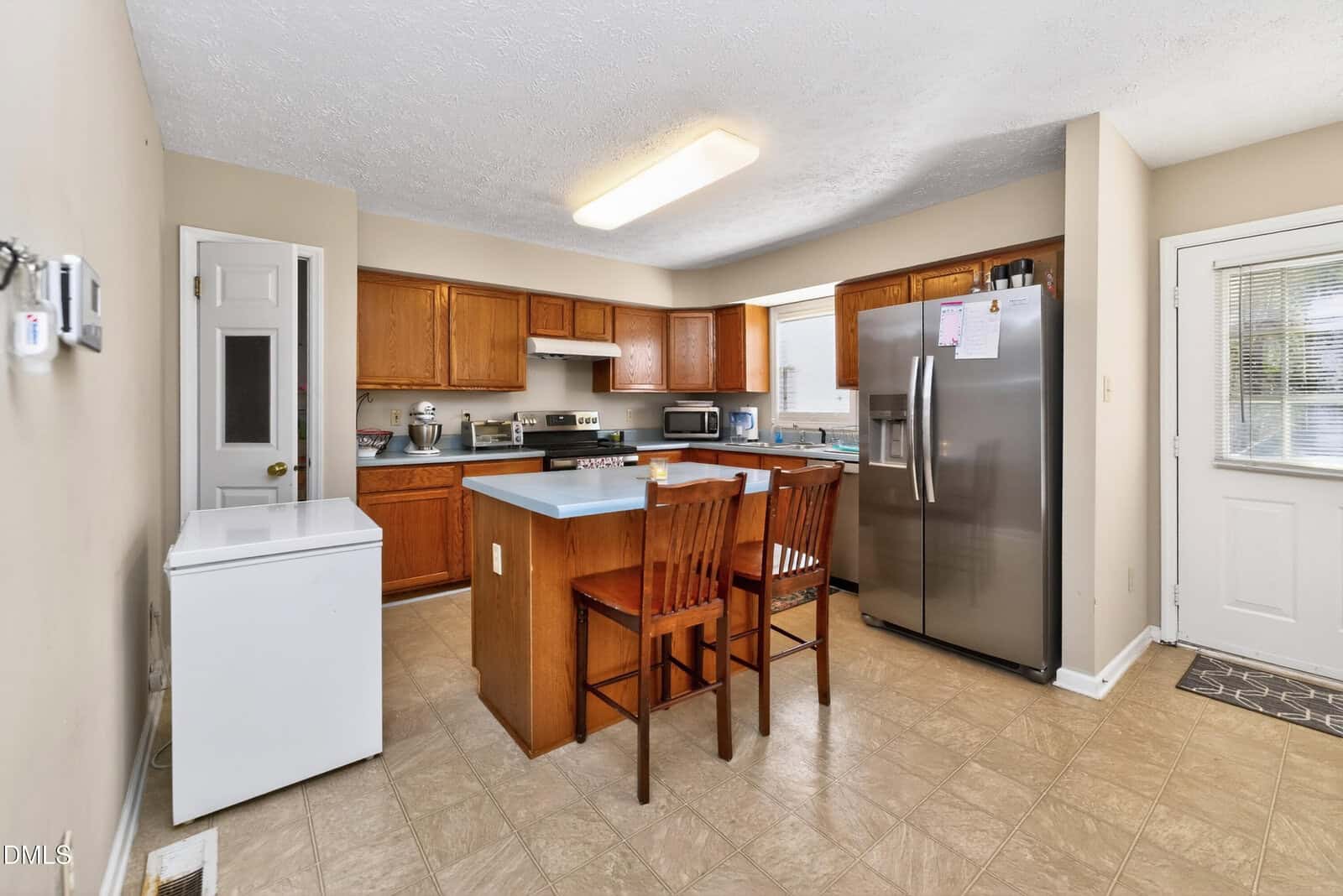 A kitchen with wooden cabinets, stainless steel appliances, a central island with two wooden stools, a white chest freezer, beige walls, tile flooring, and a door with windows leading outside.
