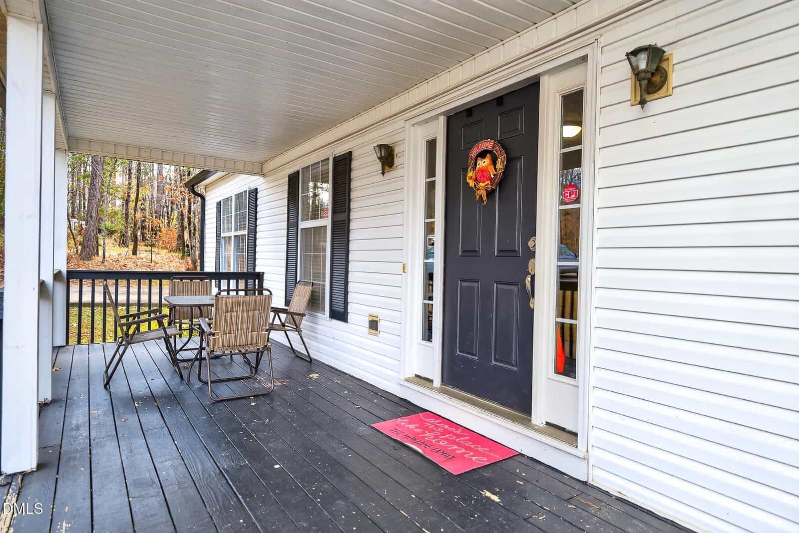 A covered front porch with black flooring, white siding, a black door with a wreath, outdoor lights, a red welcome mat, a table with four chairs, and black railings; trees are visible in the background.