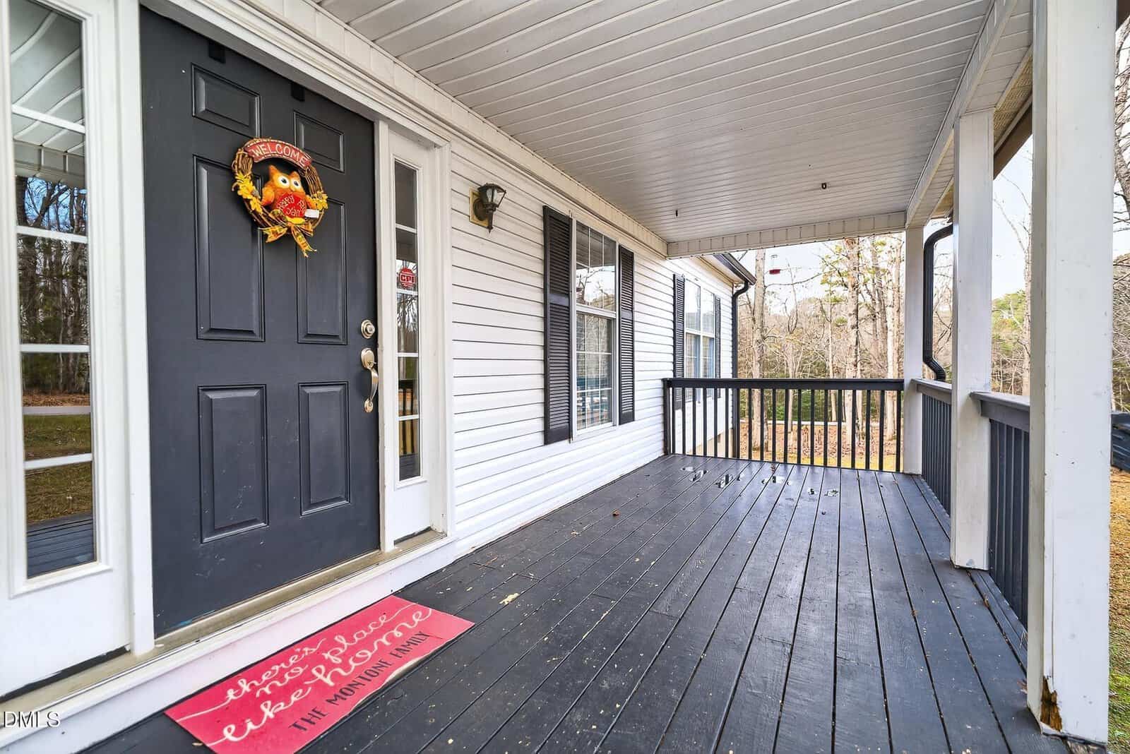 A covered front porch with black wooden flooring, white siding, and a black door decorated with an autumn wreath. A red doormat near the door reads, "There's no place like home." Trees are visible in the background.