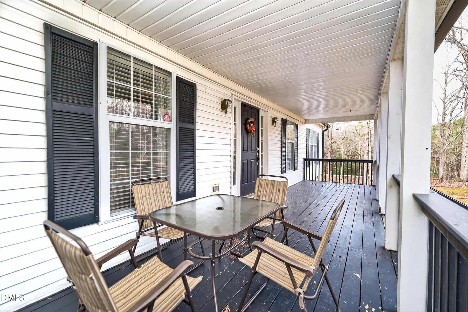 A covered porch with dark wood flooring, white siding, black shutters, and a black door. There is a glass-top table with four striped chairs and a wooded area visible in the background.