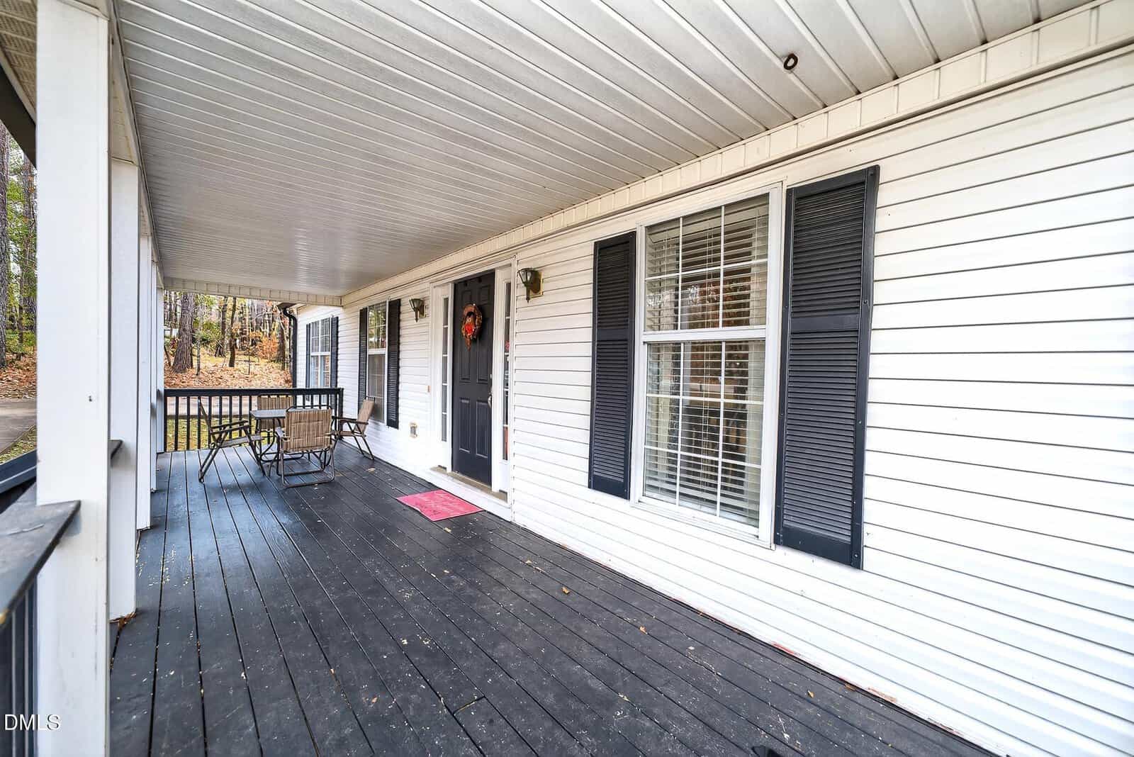 A spacious covered porch with black wooden flooring, white siding, black shutters, a black front door with a wreath, a pink doormat, and a patio table with chairs. Trees and yard are visible in the background.