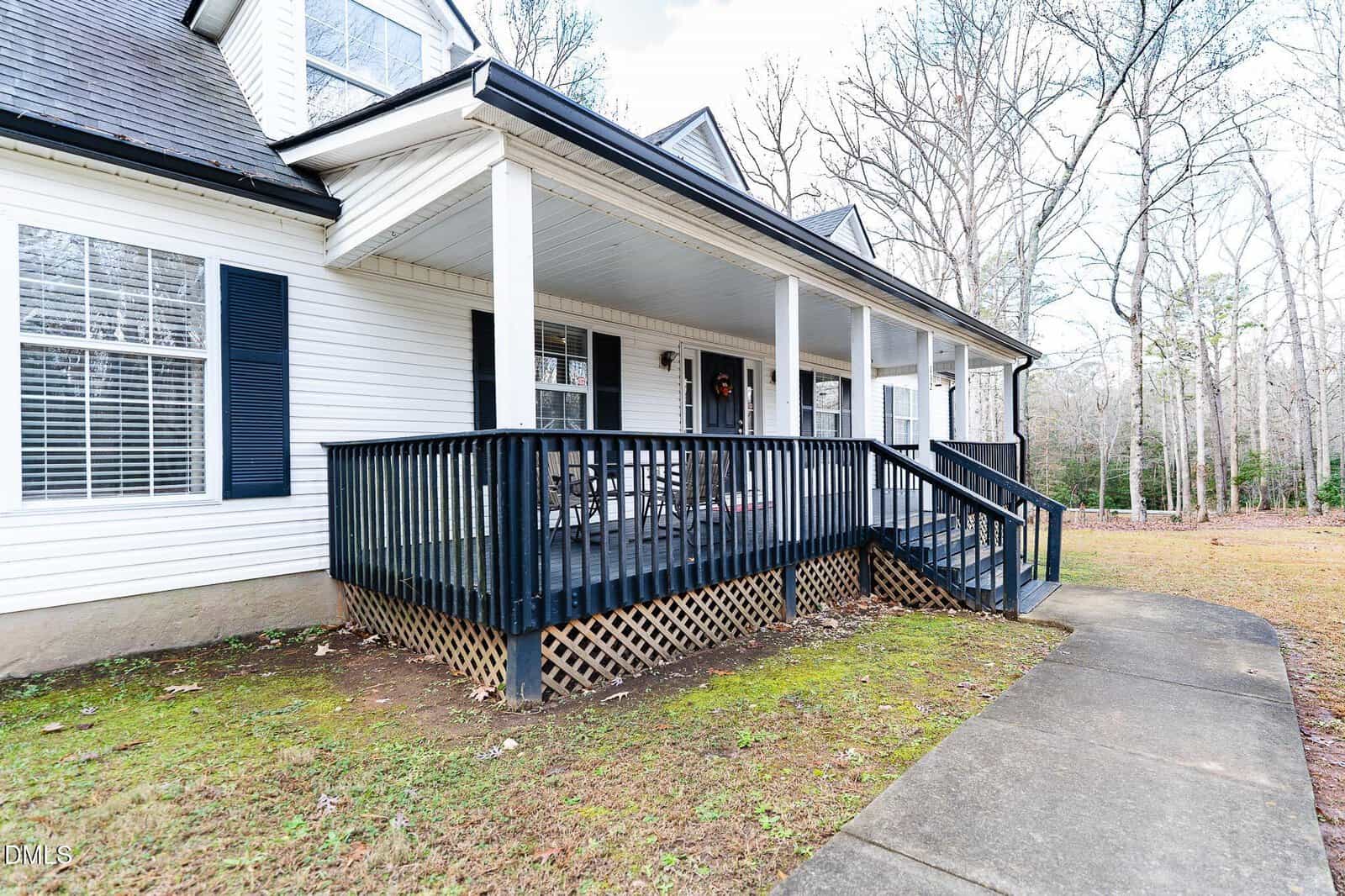 A white house with black shutters and a covered front porch featuring black railings and steps. The porch overlooks a sidewalk and a grassy yard with trees in the background.