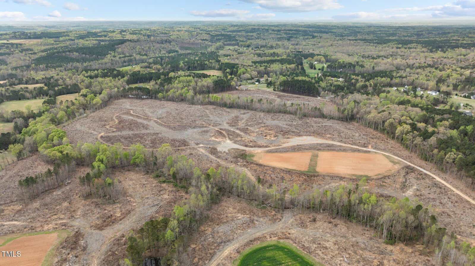 Aerial view of a large, cleared area of land surrounded by trees and forest, with patches of bare soil and winding dirt paths. Distant buildings and green fields are visible in the background under a partly cloudy sky.