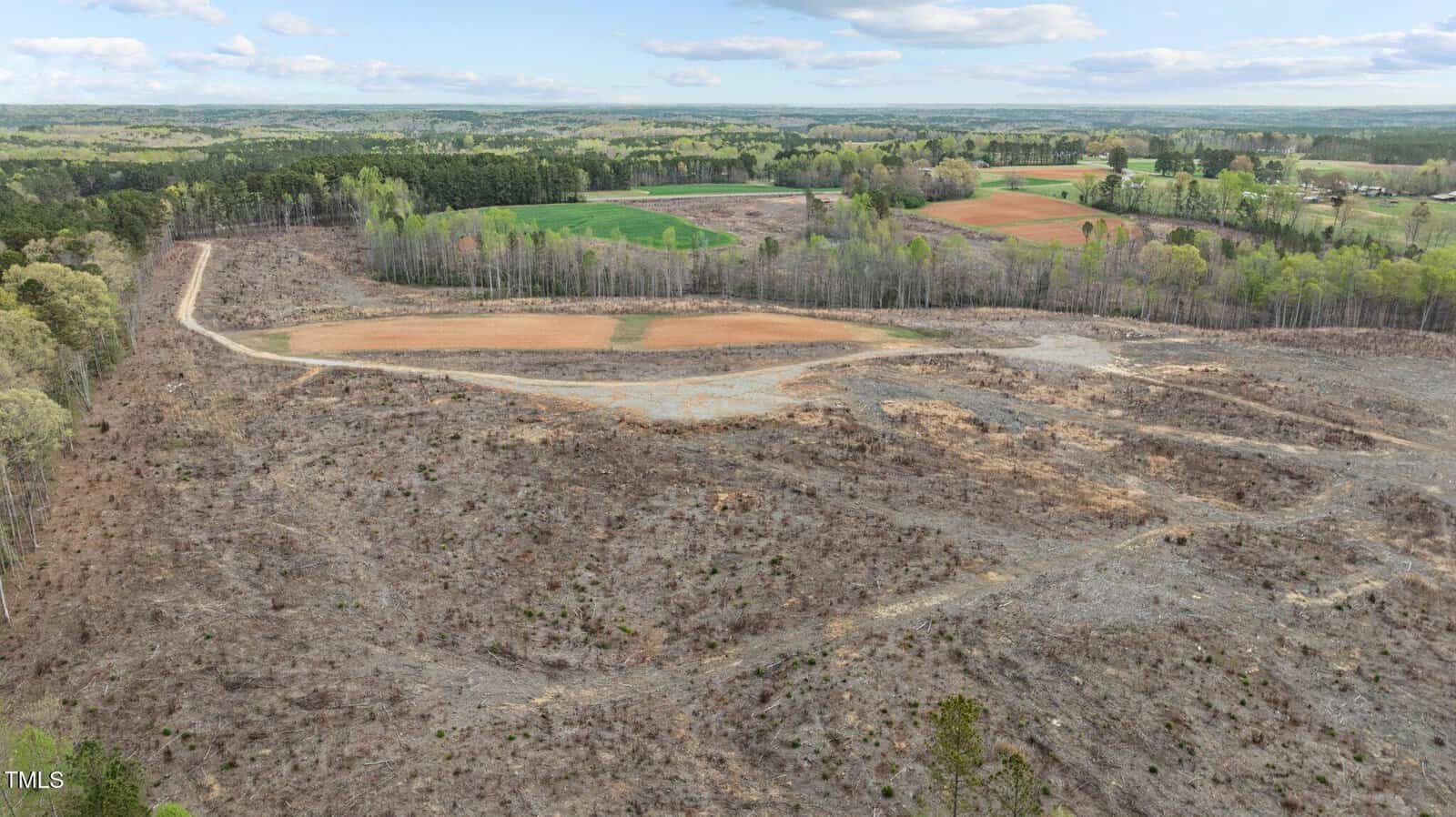 A landscape view of a partially cleared, barren field with dirt paths, surrounded by patches of trees and distant green farmland under a partly cloudy sky.